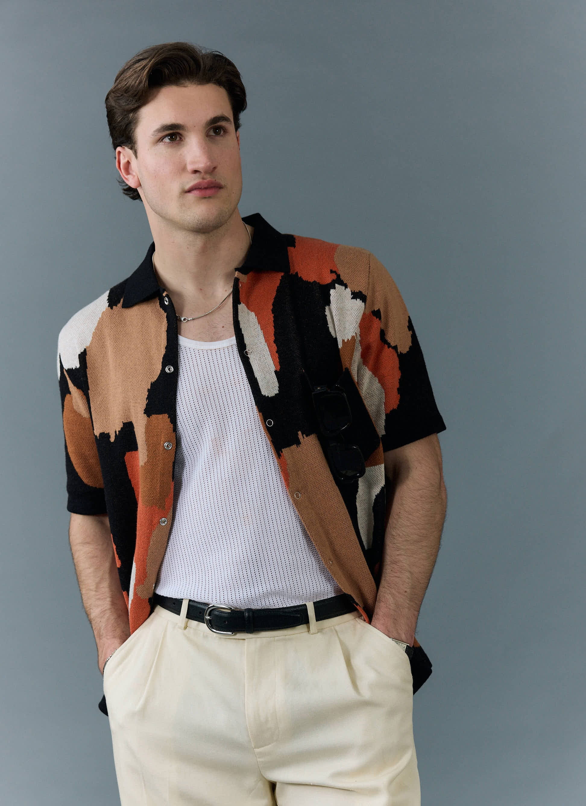 A male model wears the Percival Impressionist Jacquard Shirt in black, styled open over a white vest with cream trousers.