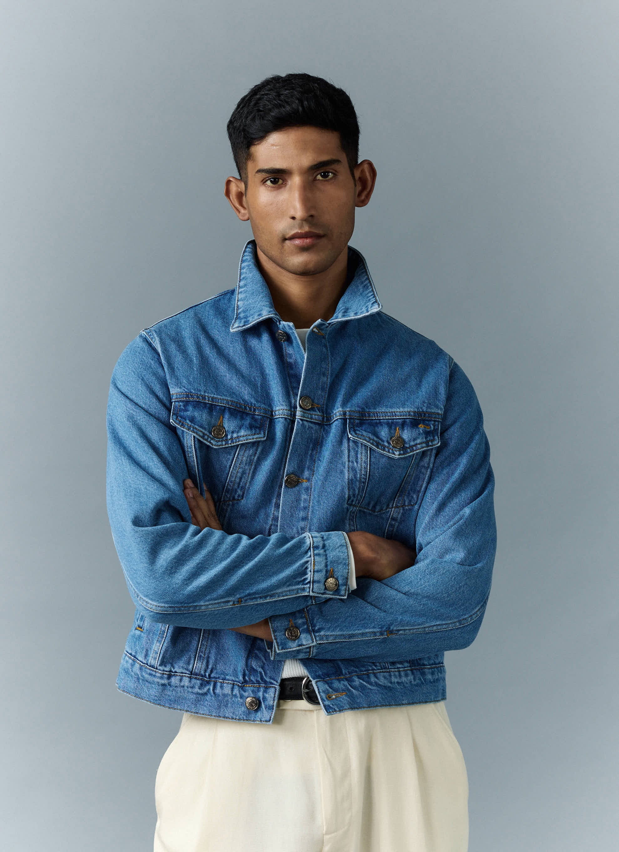 A model with arms crossed wears the light wash blue denim Western Trucker Jacket from Percival with cream trousers.