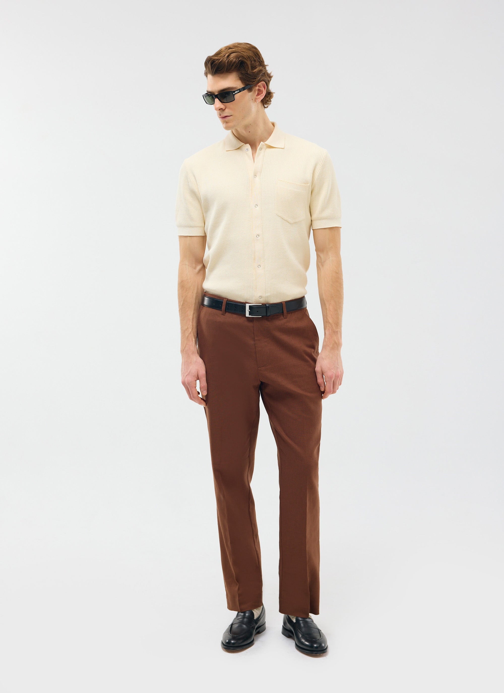 Percival Tailored Linen Trousers in brown, styled on a model with a cream knitted polo shirt, black loafers, and sunglasses.