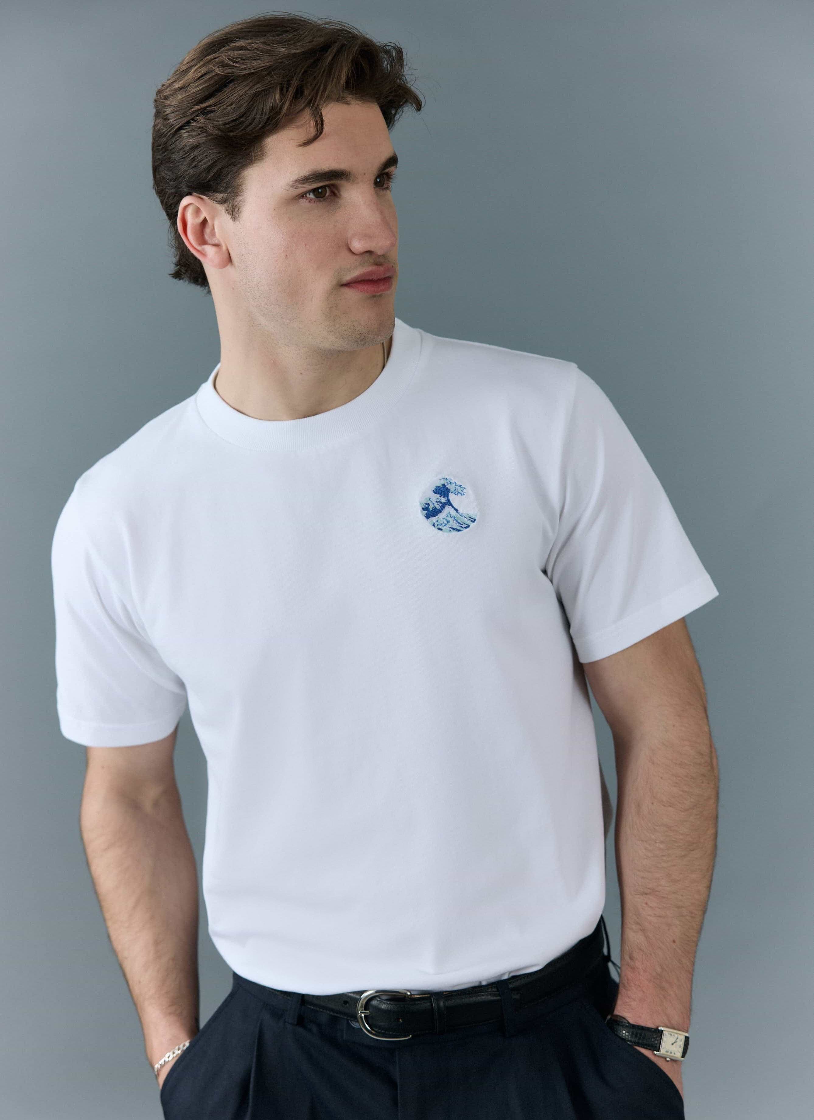 A model wears the white Percival Wave T Shirt, which features a blue and white embroidered wave design on the chest.