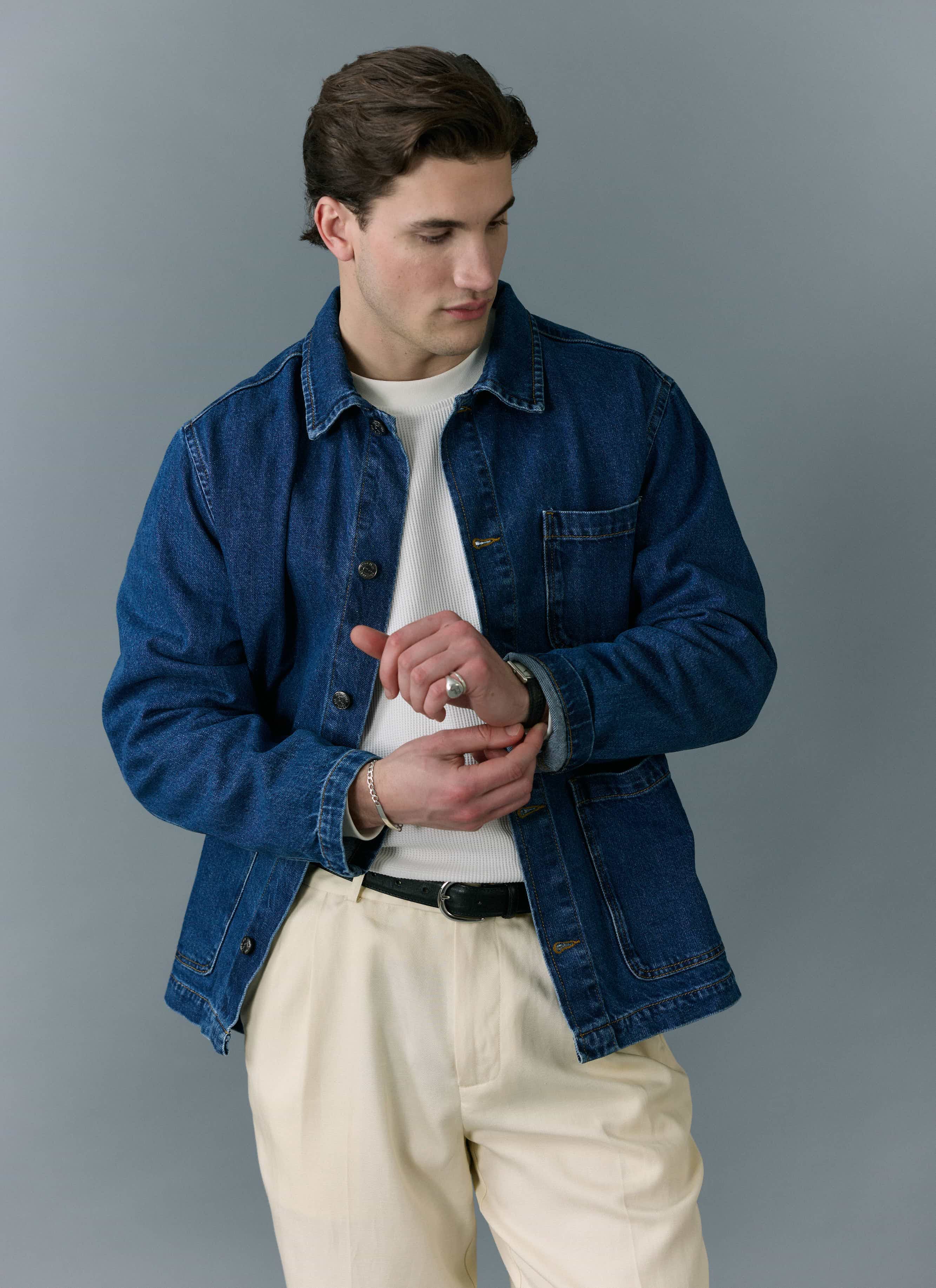A model wears the Percival Gove Chore Jacket in mid-wash blue denim, adjusting his cuff against a grey background.