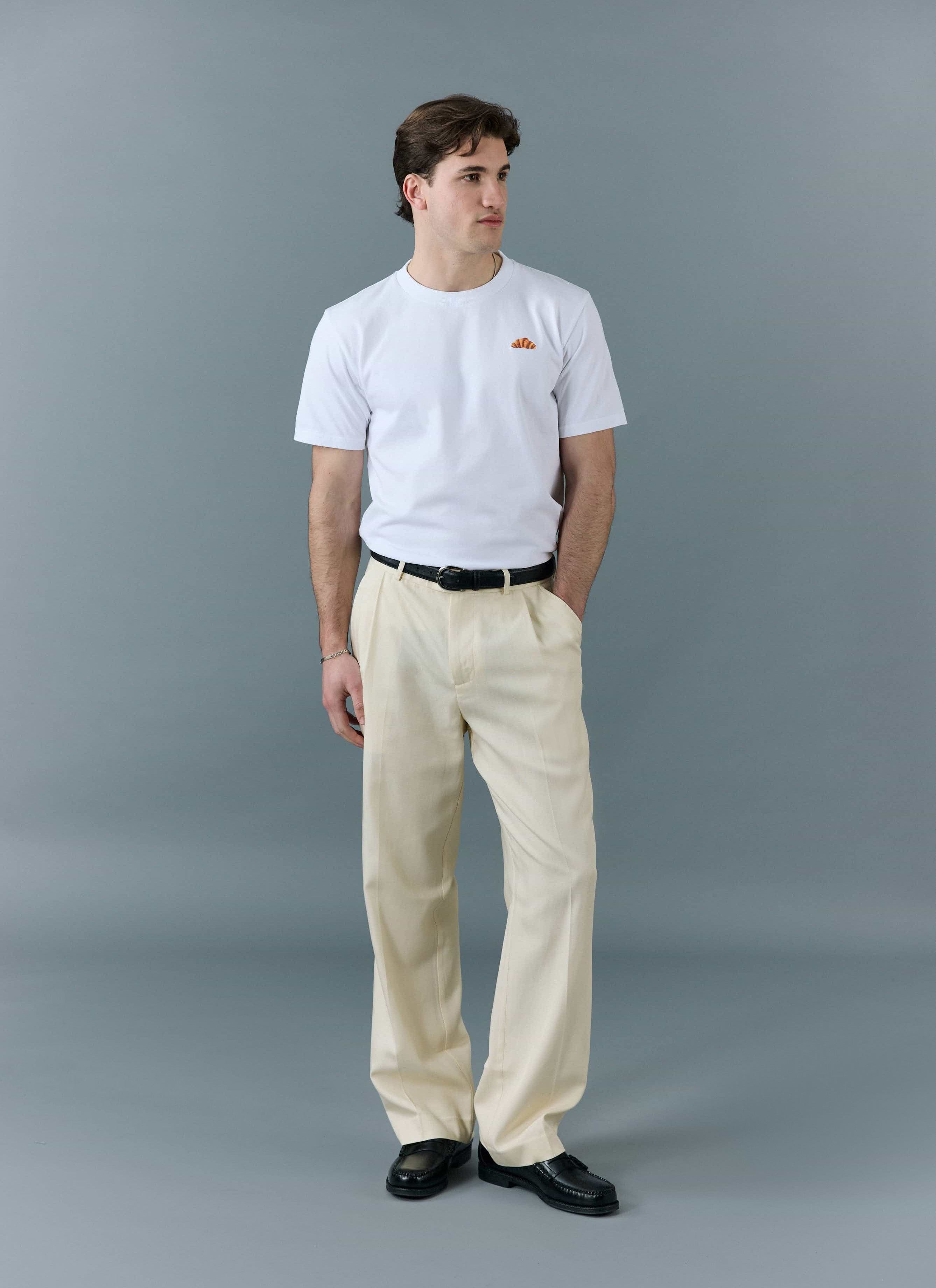 A model wears the Percival white Croissant T-Shirt, tucked into cream trousers with a black belt, against a grey background.
