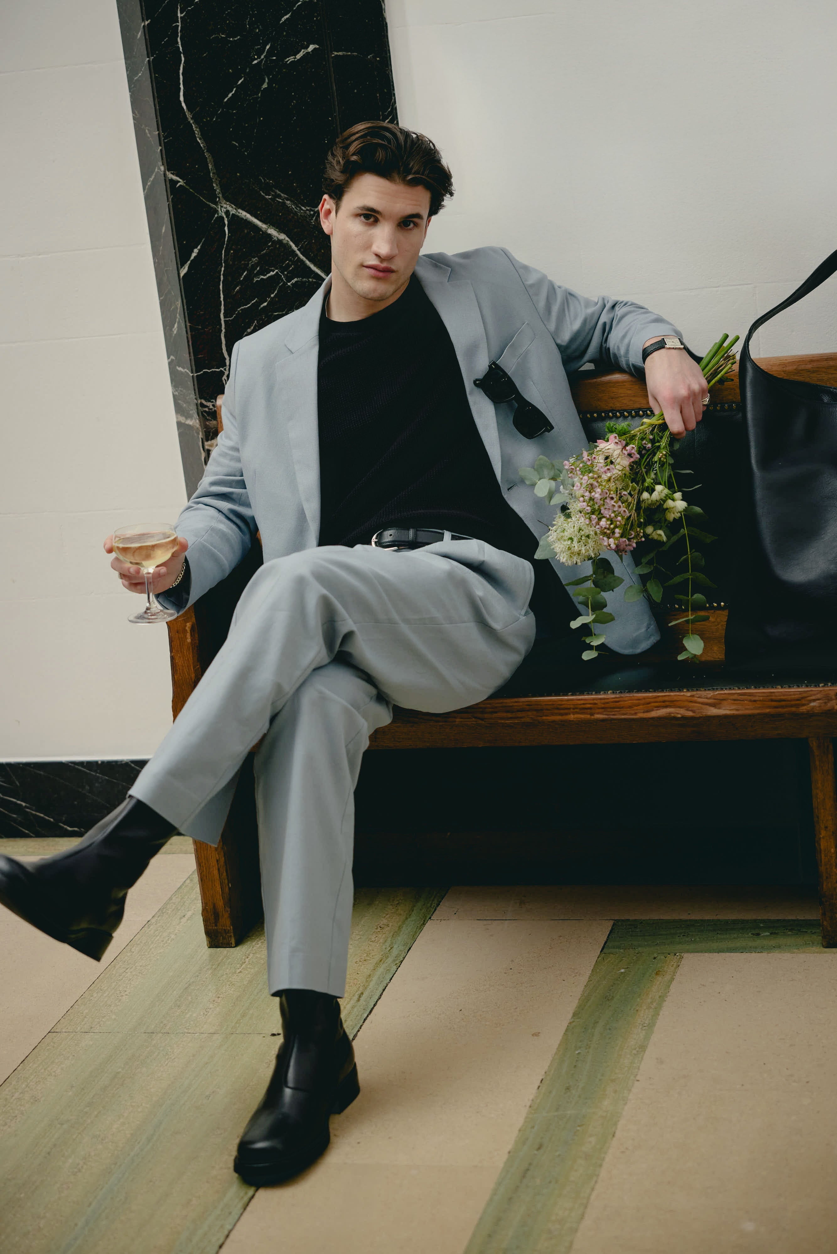 A model sits on a bench wearing the Percival light blue linen suit, styled with a black jumper, holding flowers.