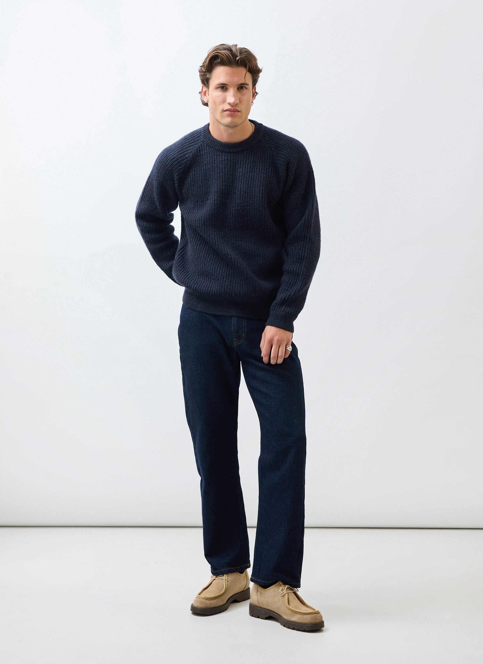 A man models the Percival Fisherman Raglan Jumper in navy wool, styled with dark jeans against a white background.