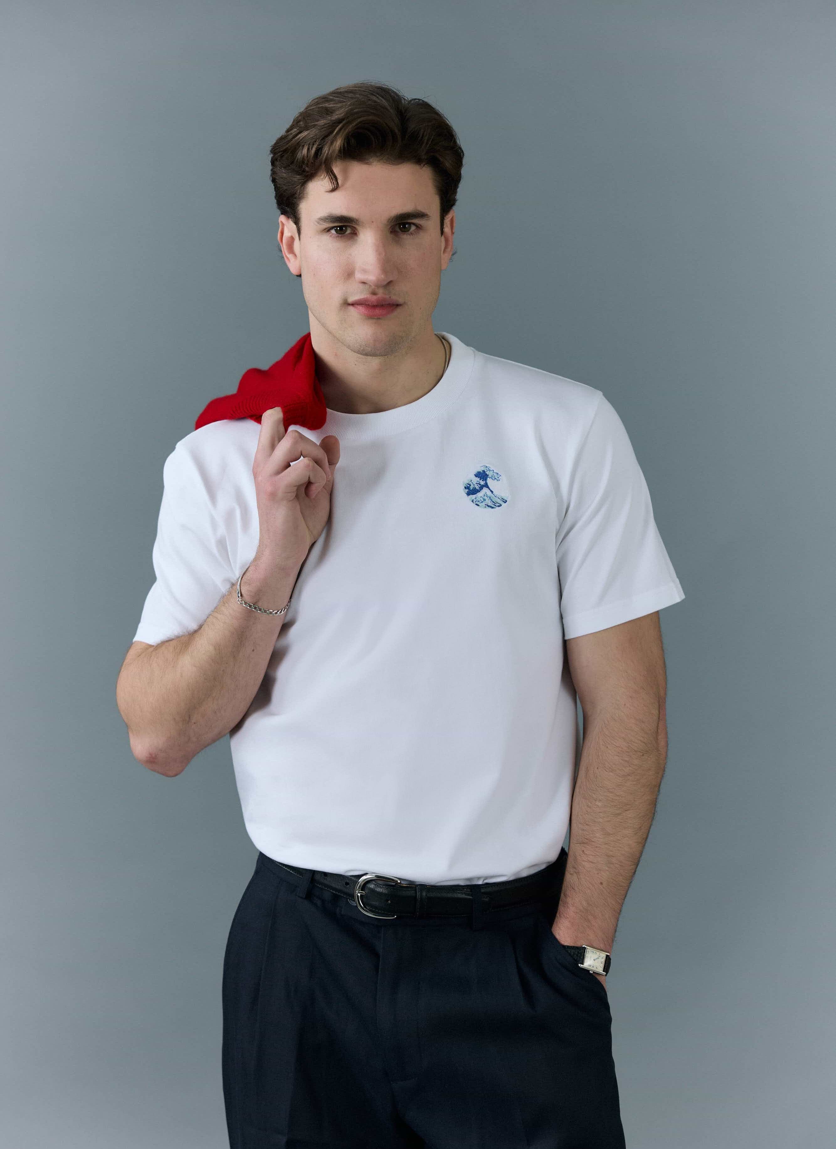 A model wears the white Percival Wave T Shirt with blue embroidery, styled with dark trousers and a red jumper on shoulder.