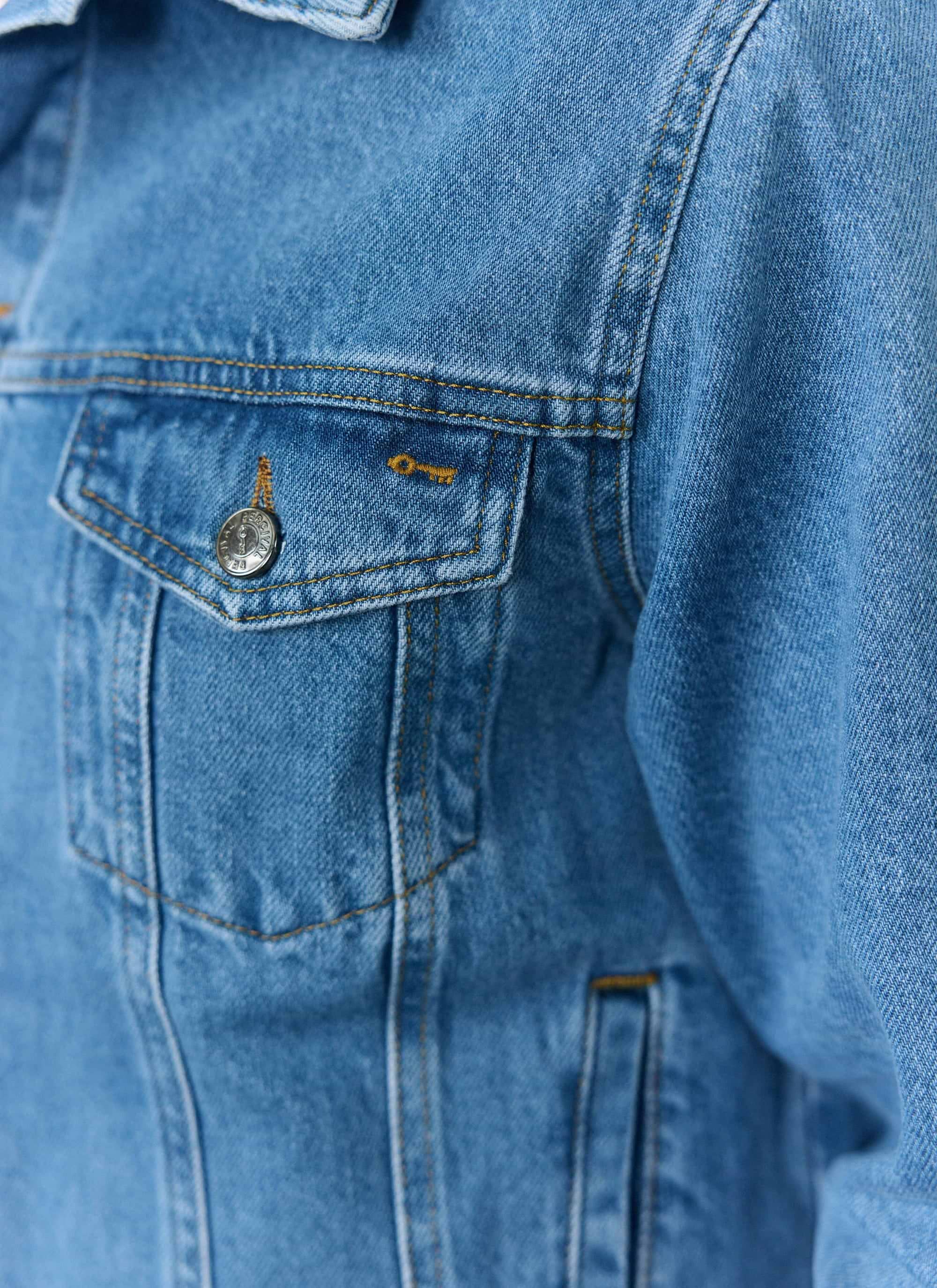 A close-up of the Percival Western Trucker Jacket in light wash blue denim, showing the chest pocket and branded button.