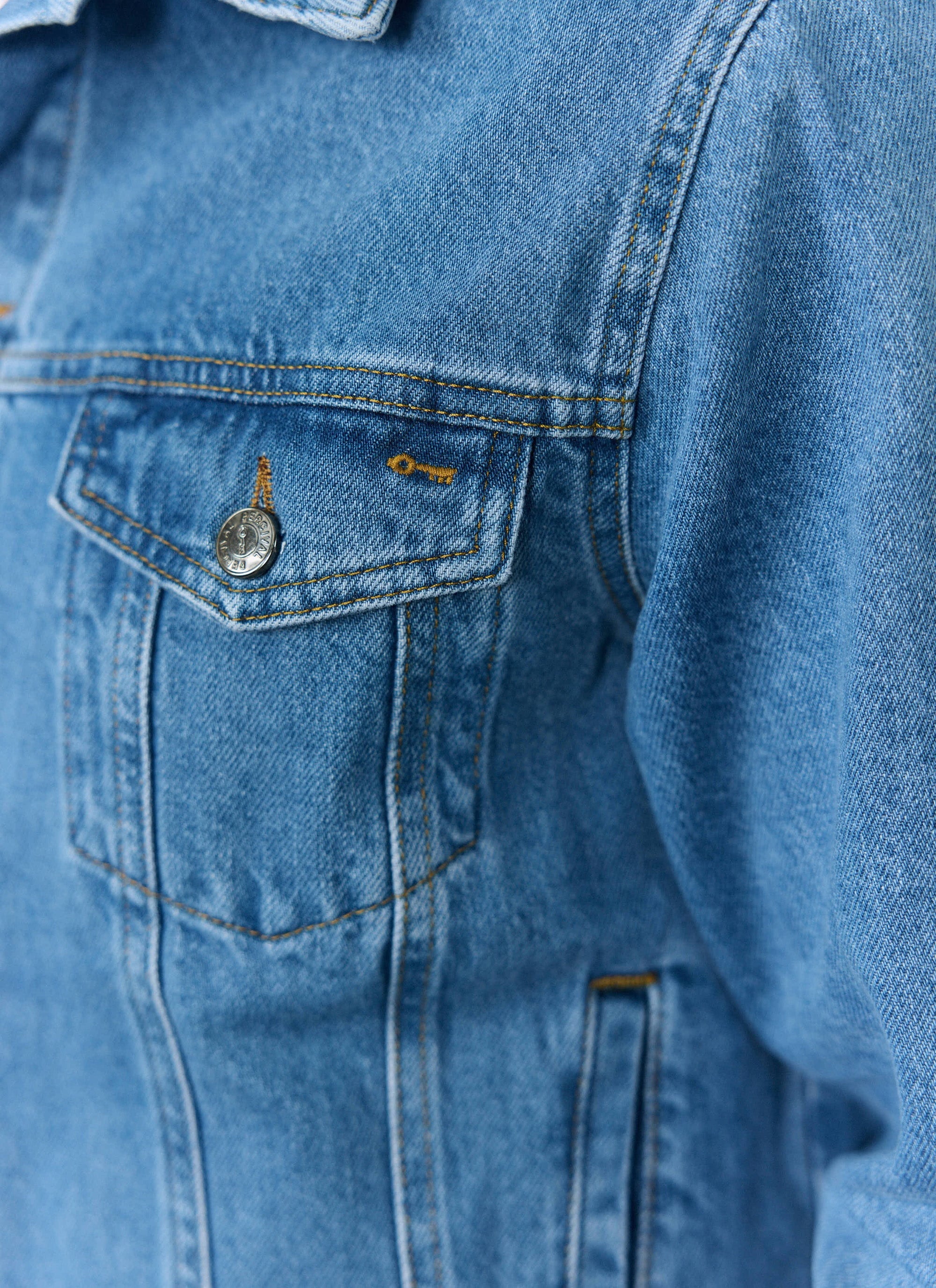 A close-up of the Percival Western Trucker Jacket in light wash blue denim, showing the chest pocket and branded button.