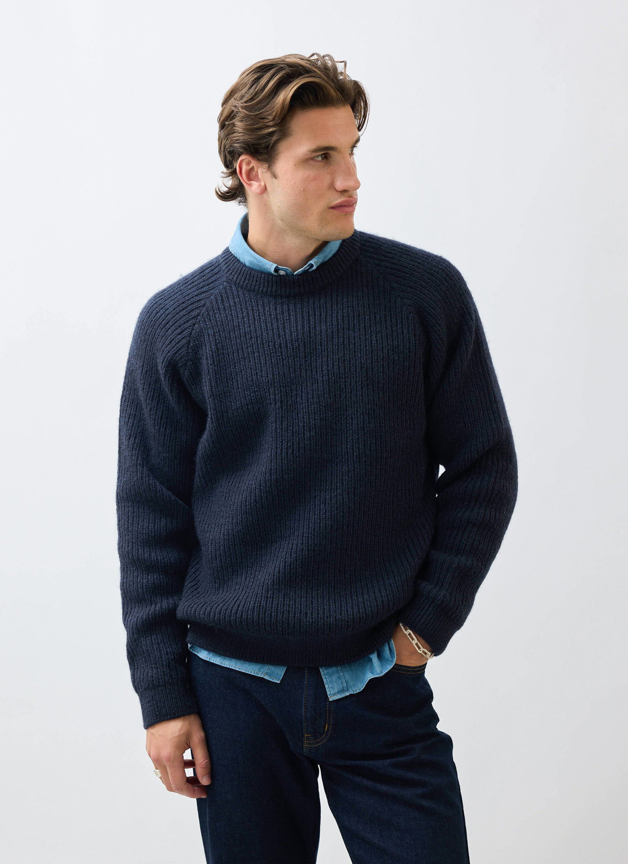 A man models the Percival Fisherman Raglan Jumper in navy wool, layered over a denim shirt and paired with dark jeans.