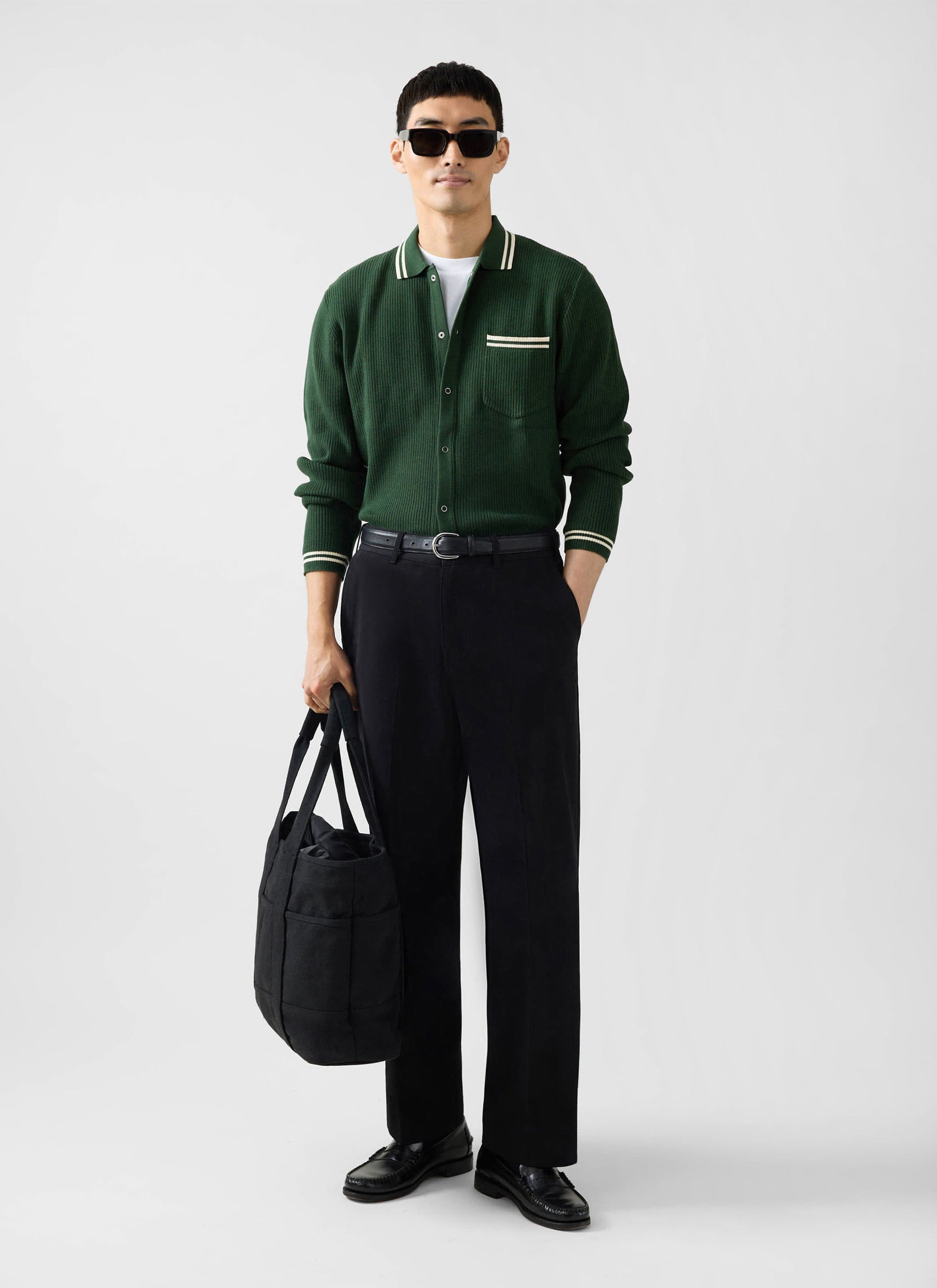 A model wears the Percival Pablo cotton knitted shirt in forest green with black trousers and sunglasses.