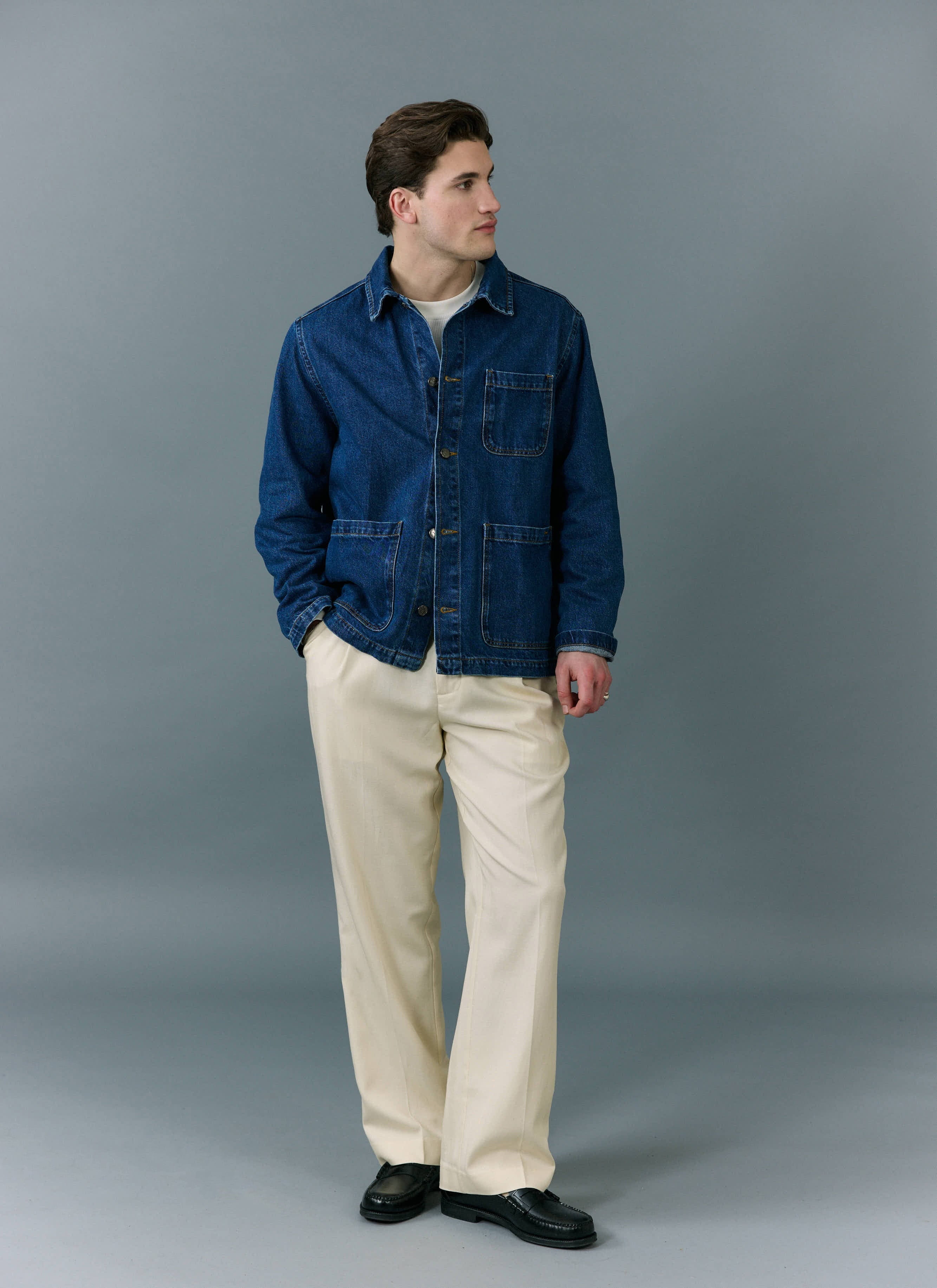 A man models the Percival Gove Chore Jacket in mid wash blue denim, styled with cream trousers and black loafers.
