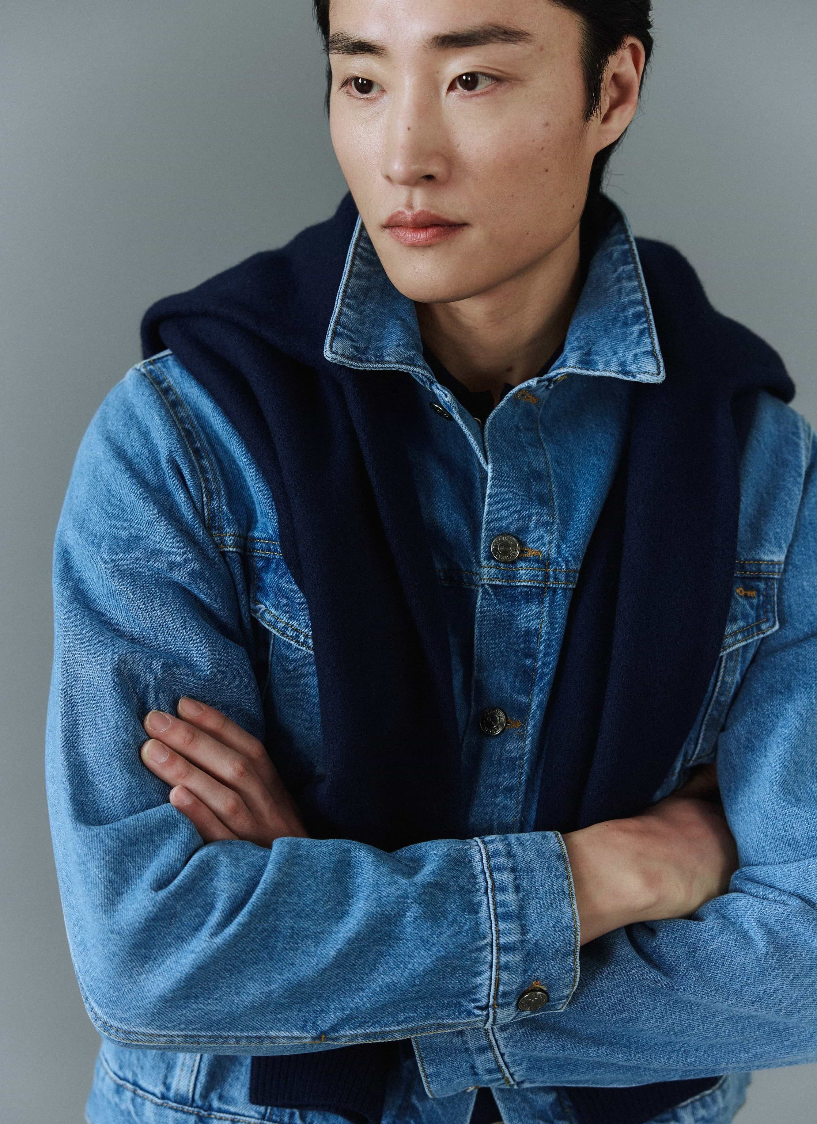 A model wears the Percival Western Trucker Jacket in light wash blue denim with a navy knit layered over his shoulders.