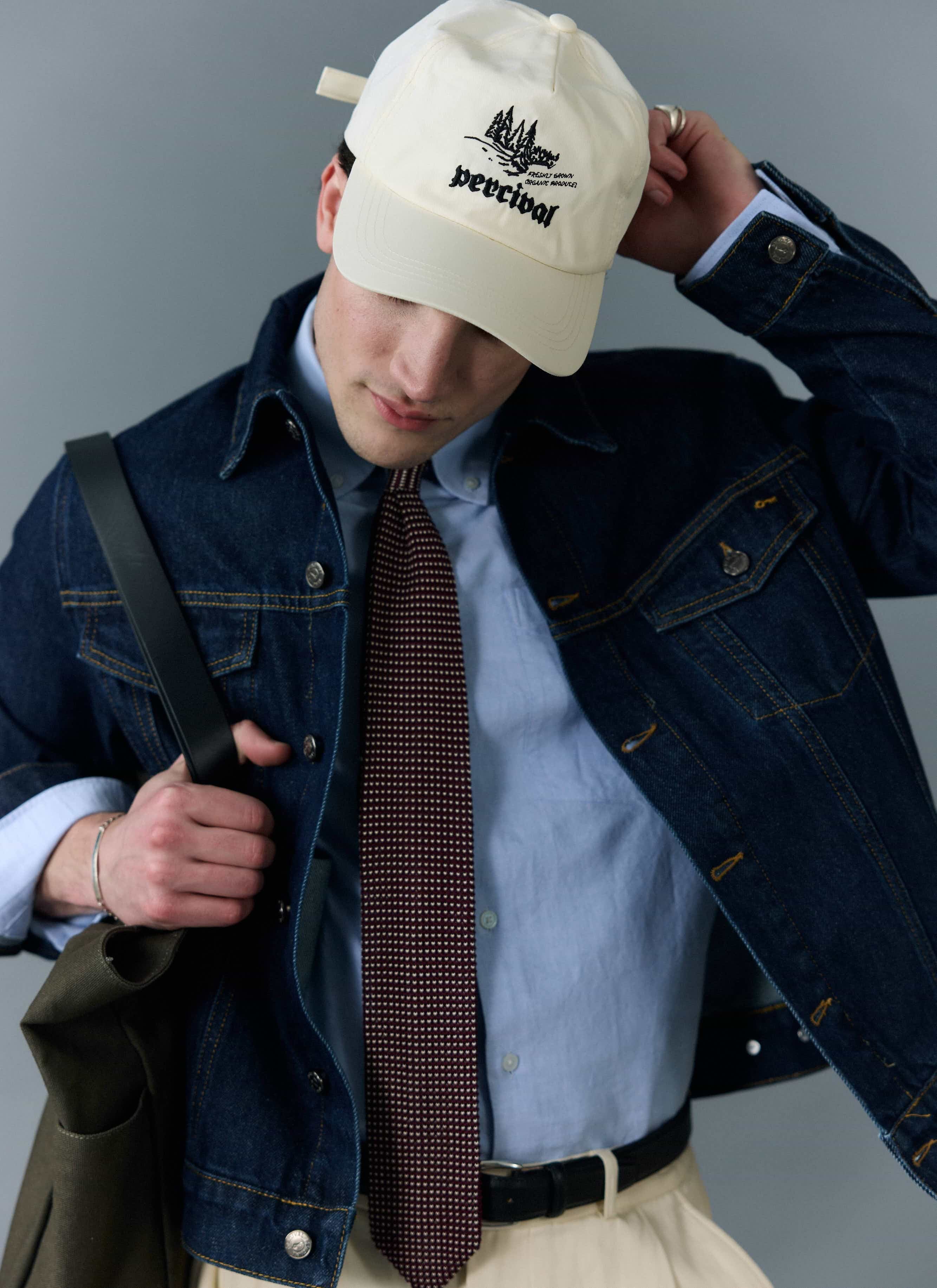 A man models the Percival Western Trucker Jacket in dark indigo denim, styled with a shirt, tie and baseball cap.