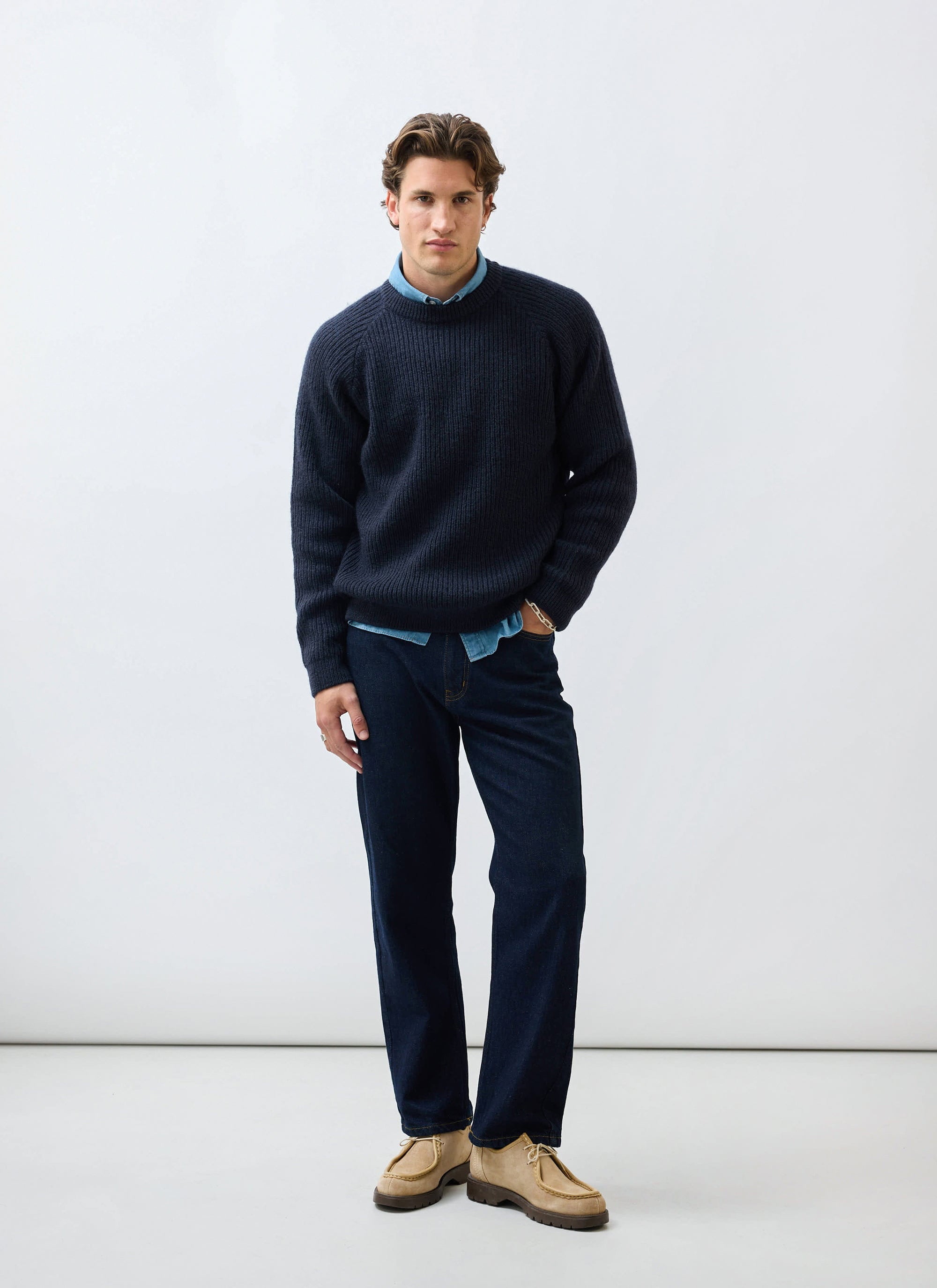A model styles the Percival Fisherman Raglan Jumper in navy wool over a blue shirt with dark denim jeans.