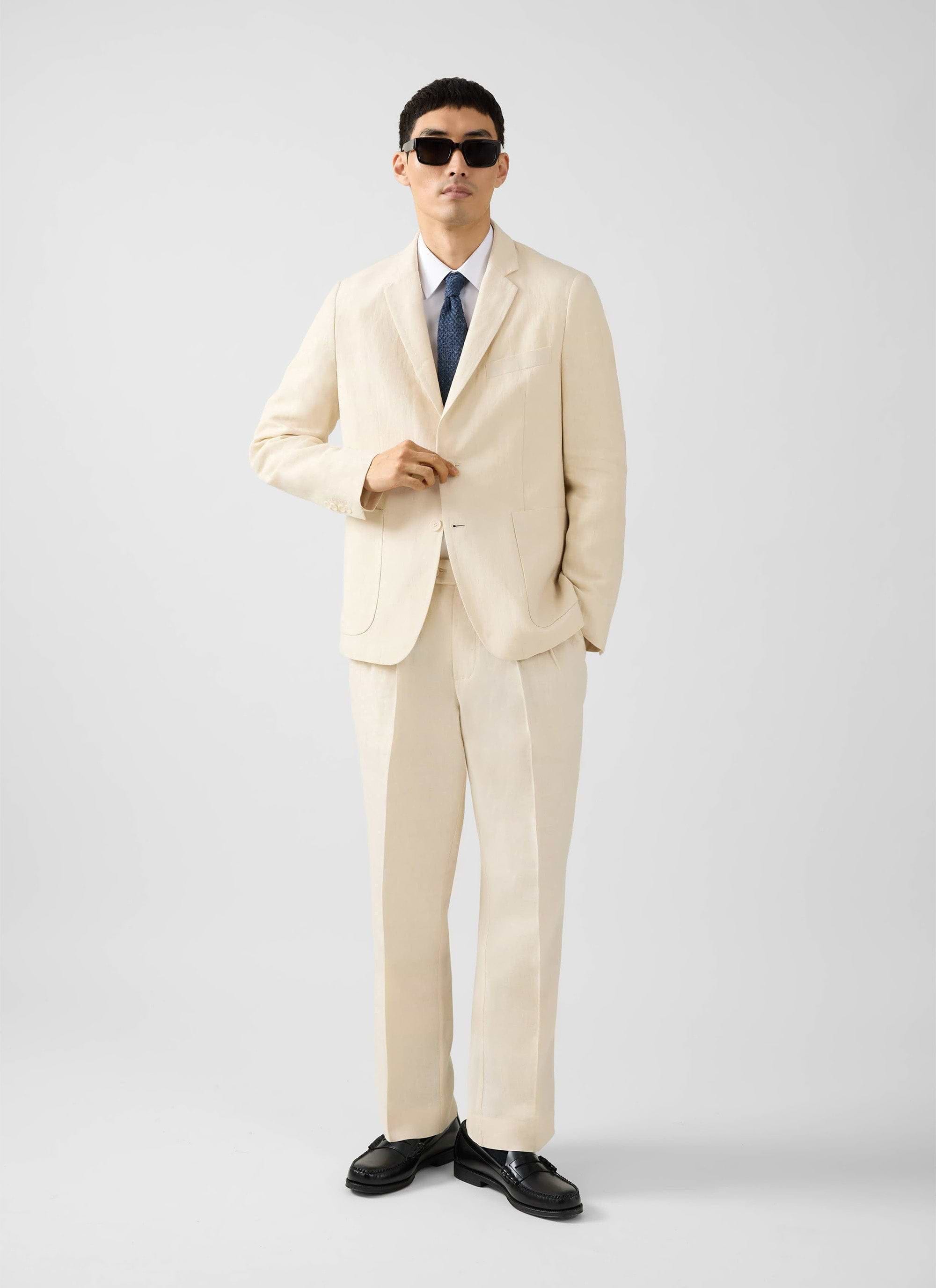 A model wears the Percival cream tailored linen blazer with matching trousers, a shirt, tie, and sunglasses.