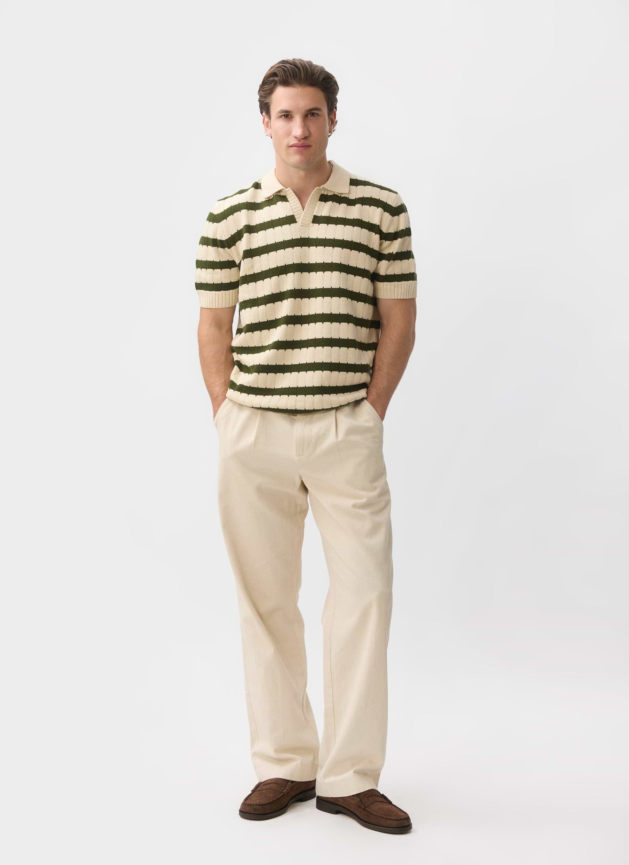 A man models the Percival Stine Knitted Polo in ecru with green stripes, with ecru trousers against a white background.