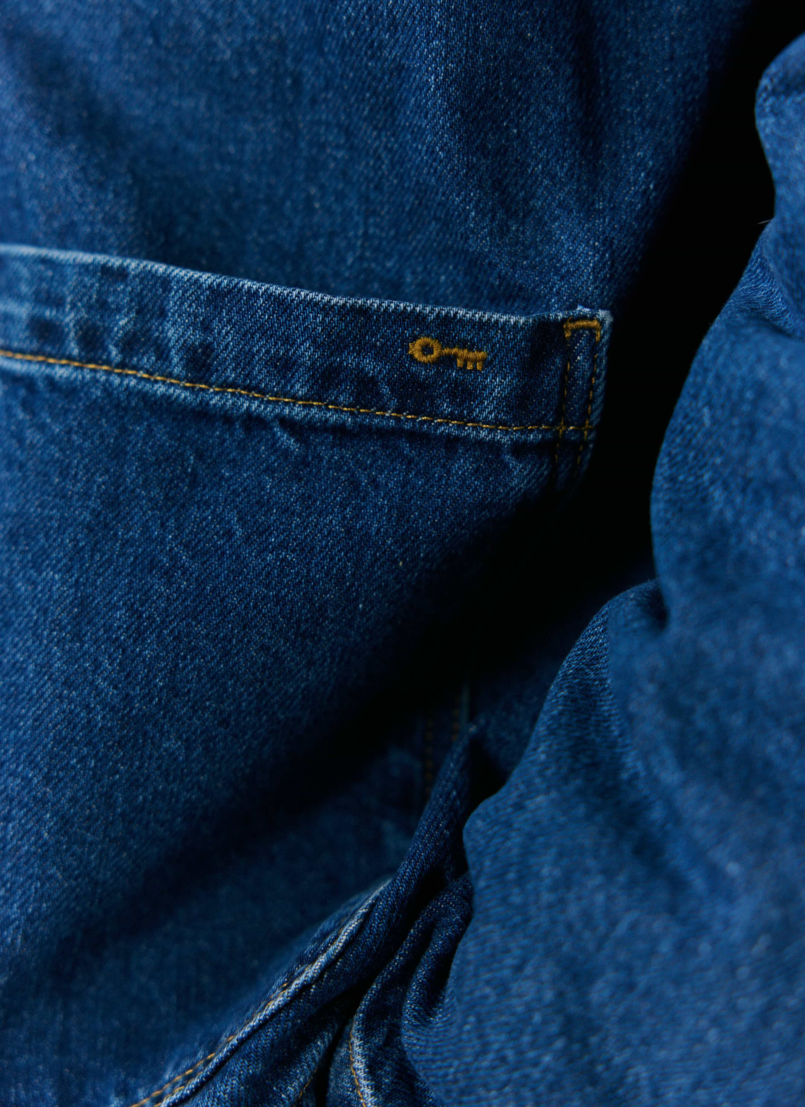 A close-up of the Percival Gove Chore Jacket in mid-wash blue denim, showing the key logo embroidery on the pocket.