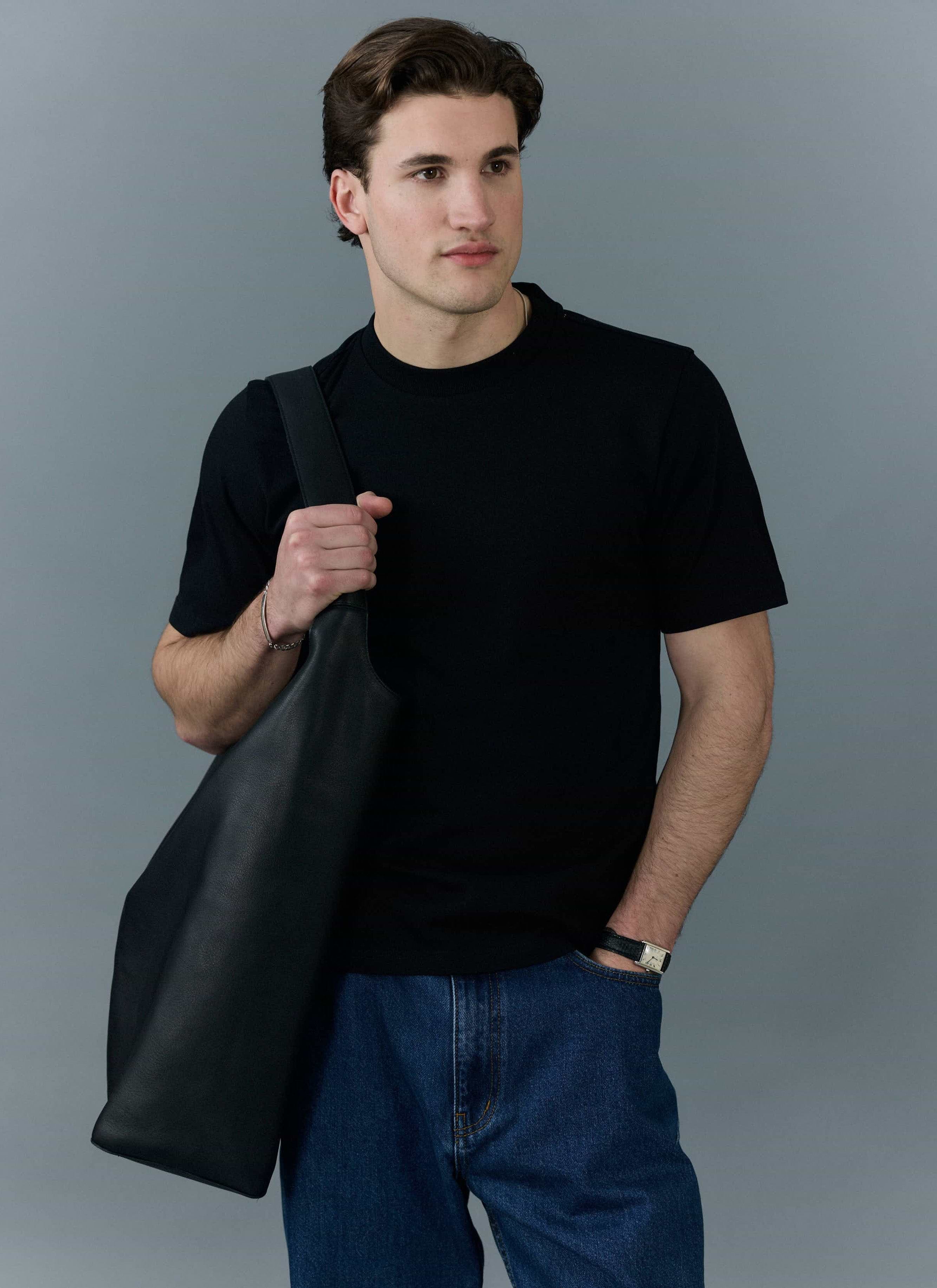 A man models the Percival Cropped Regular Fit T Shirt in black with blue jeans and a black tote bag.