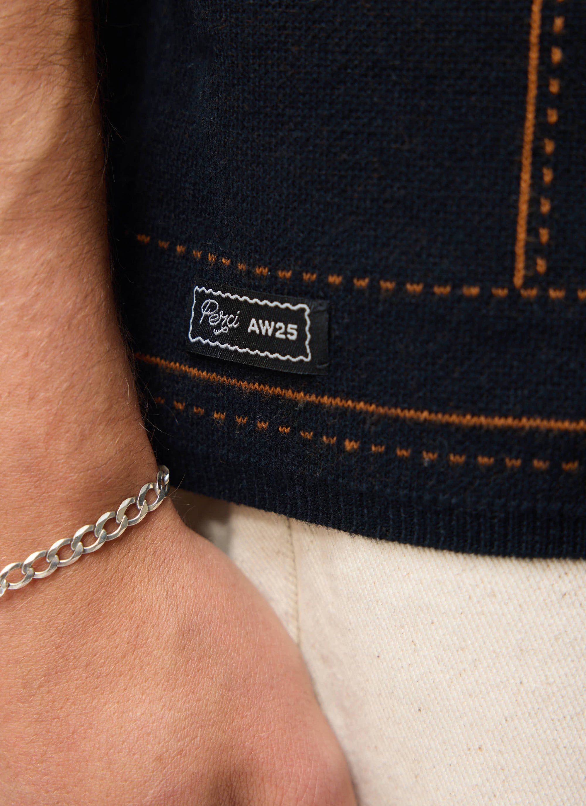 A close-up shot of the Percival Folio Knitted Shirt in dark navy, showing the jacquard detail and AW25 branded hem label.