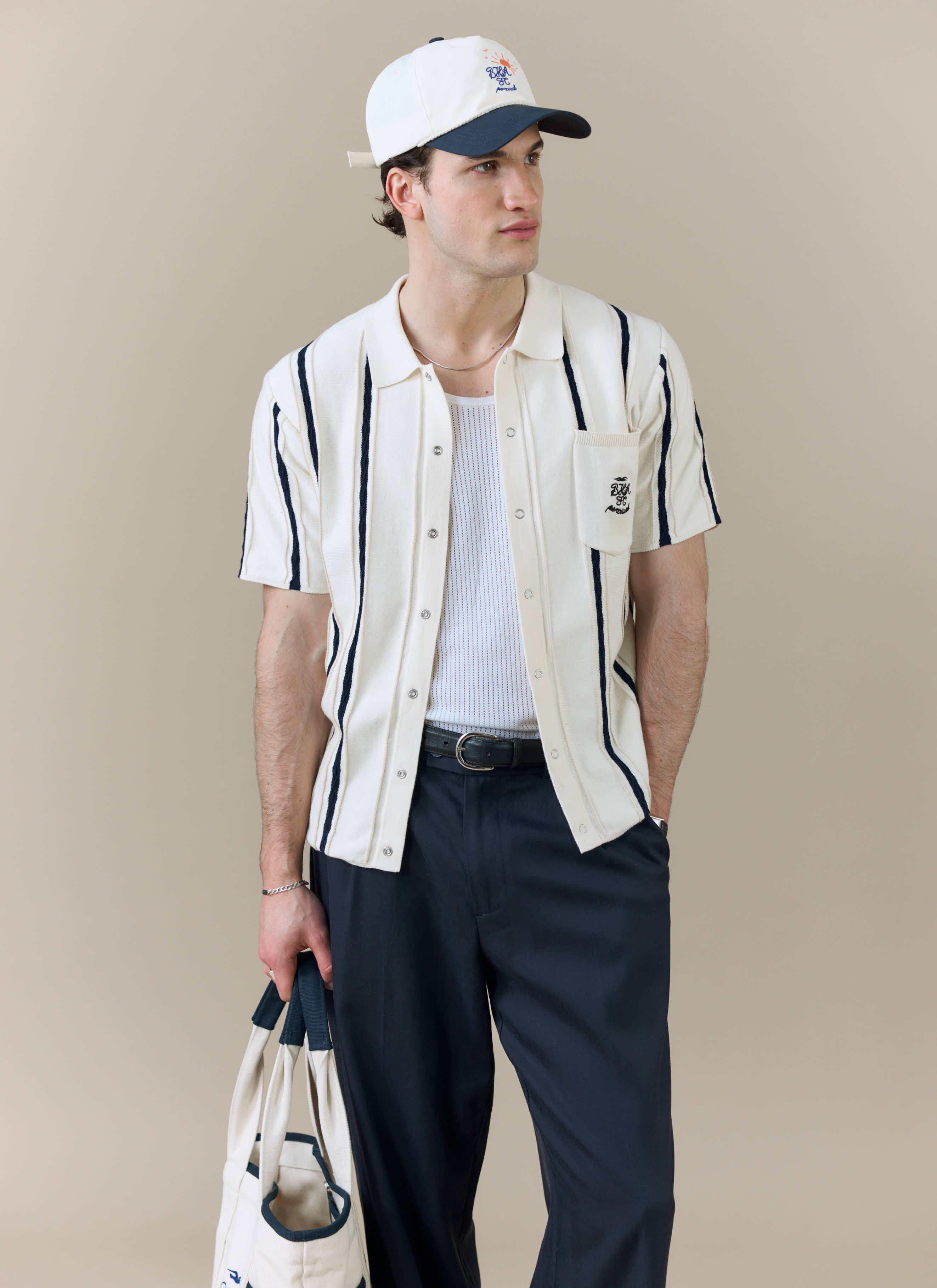 A model wears the Percival x Brighton & Hove Albion F.C. Cable Stripe Knitted Shirt in white, styled with a cap and tote bag.