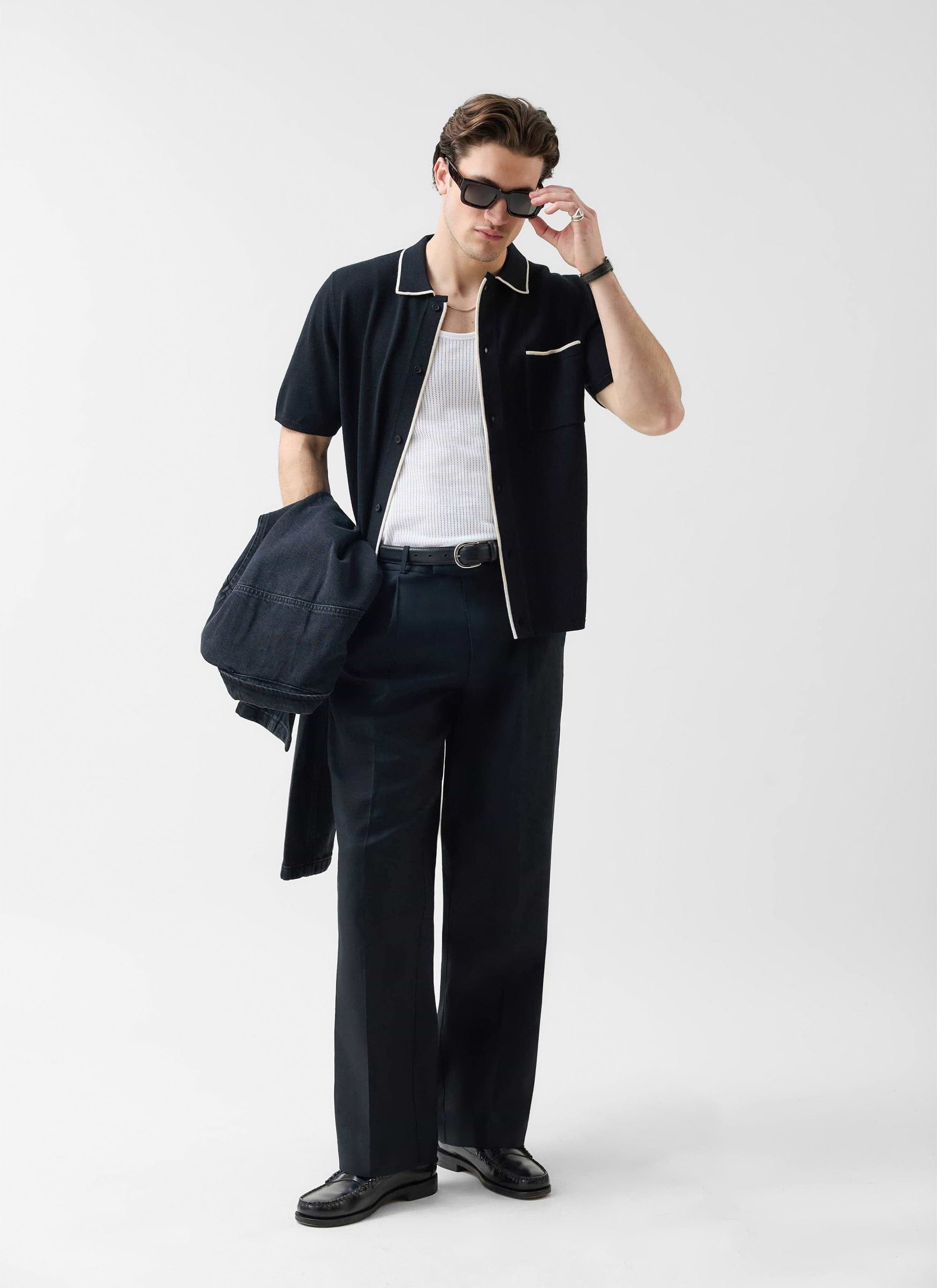 A model wears the black Percival Contrast Trim Knitted Shirt layered over a white vest with black trousers and sunglasses.
