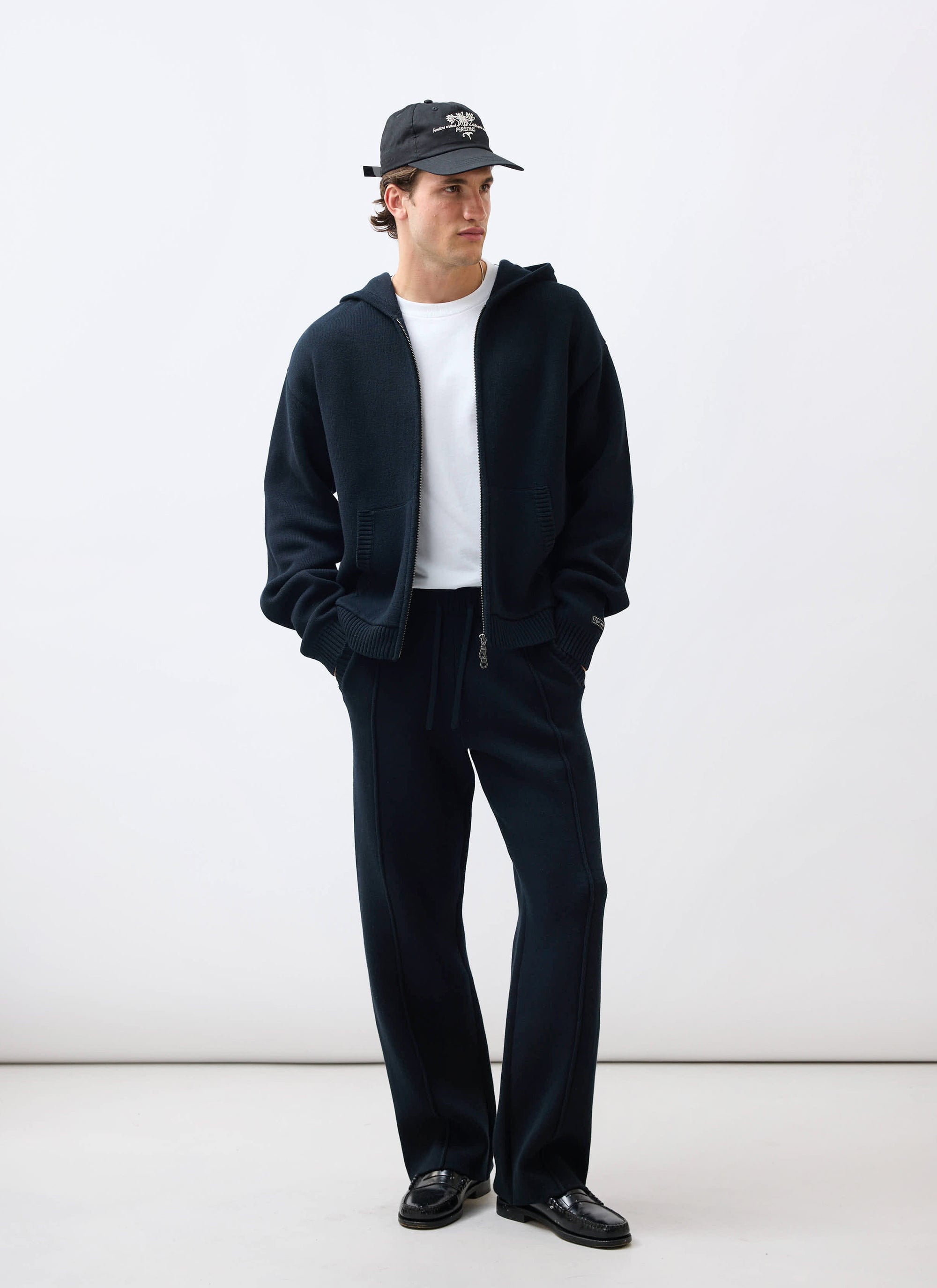 A model wears the black Percival Chronicle Knitted Hoodie and matching track pants, styled with a white tee and a cap.