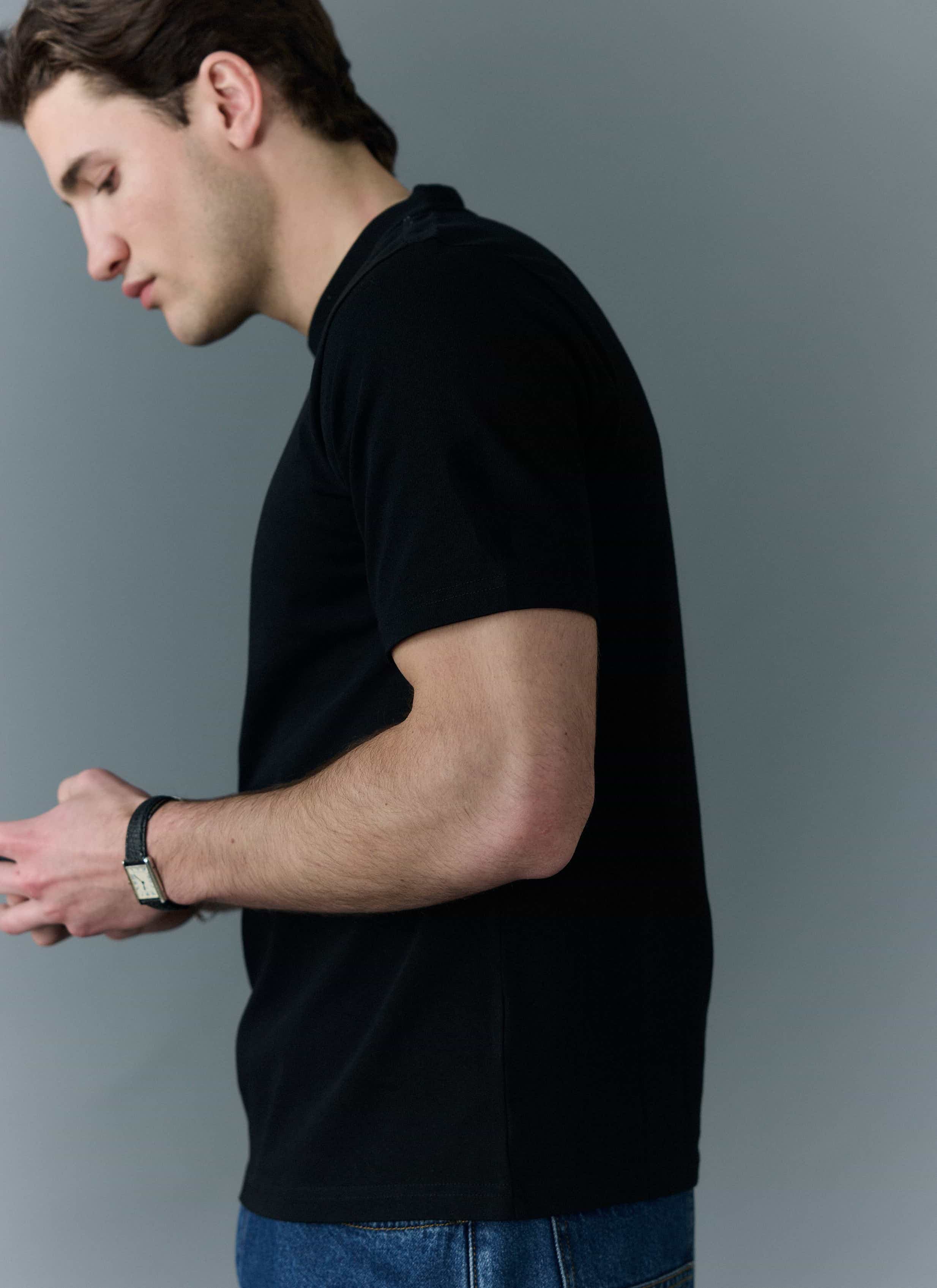 A model wears the Percival Cropped Regular Fit T-shirt in black organic cotton, viewed from the side against a grey wall.