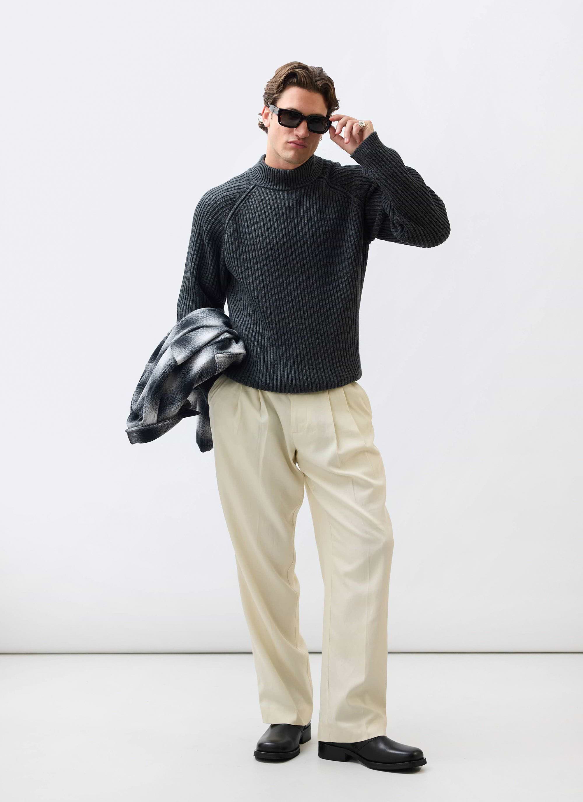 A man models the Percival Library Turtle Neck in charcoal with cream trousers, adjusting his black sunglasses.