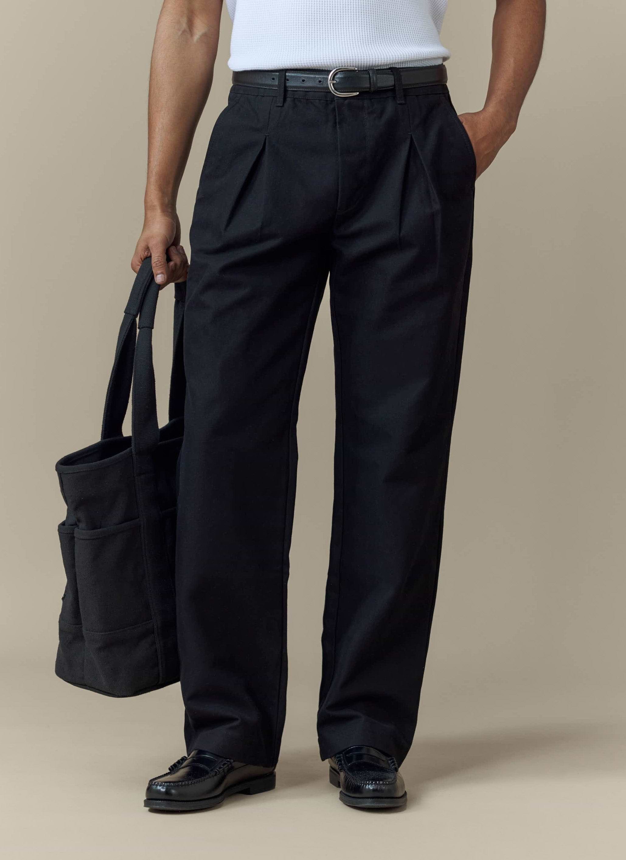 A model wears the Percival Seb Inverted Pleat Trousers in black, styled with a white vest, loafers and a tote bag.