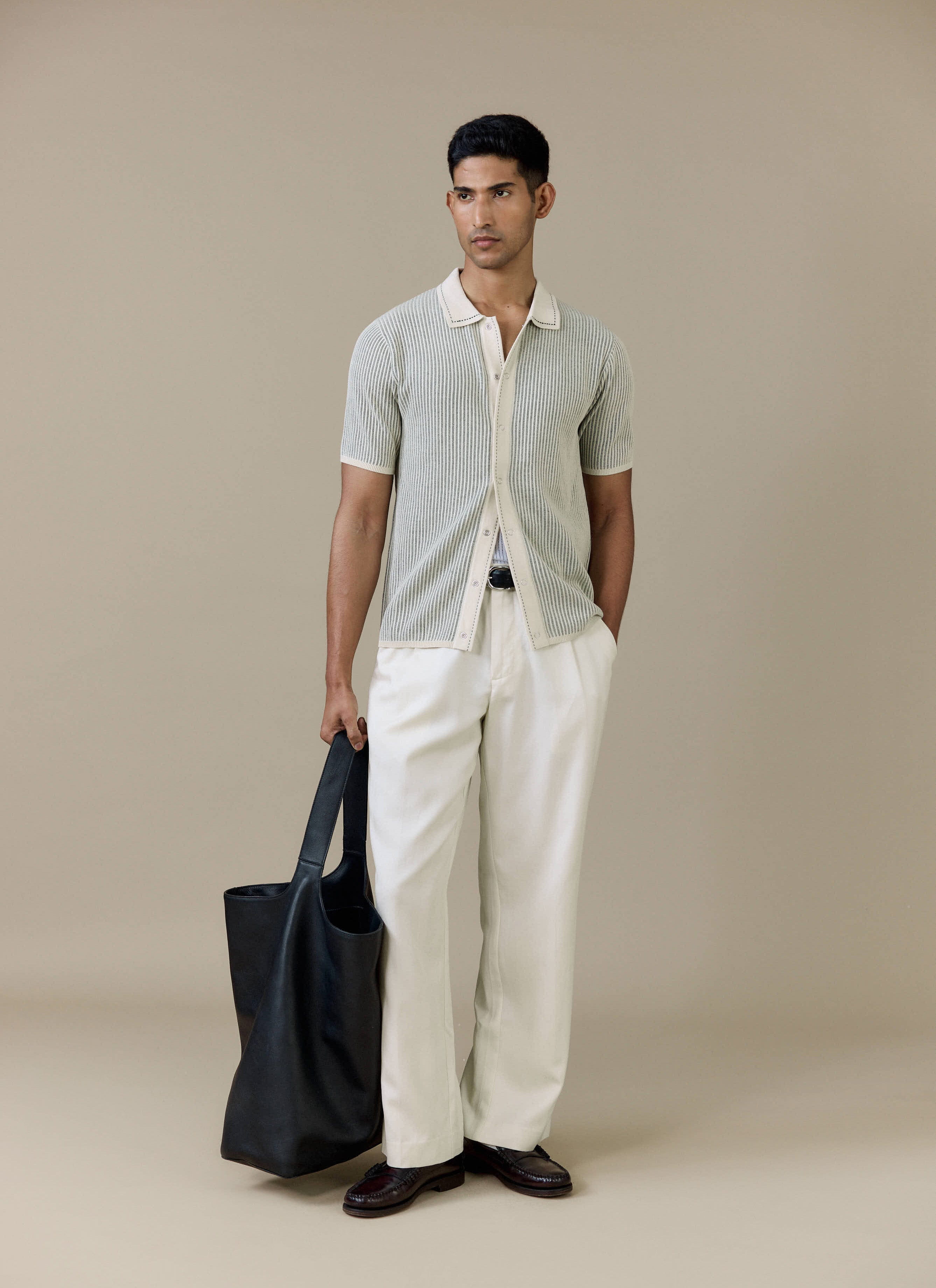 A model wearing the cream Percival Dixon Knitted Shirt with white trousers and a black leather tote bag.