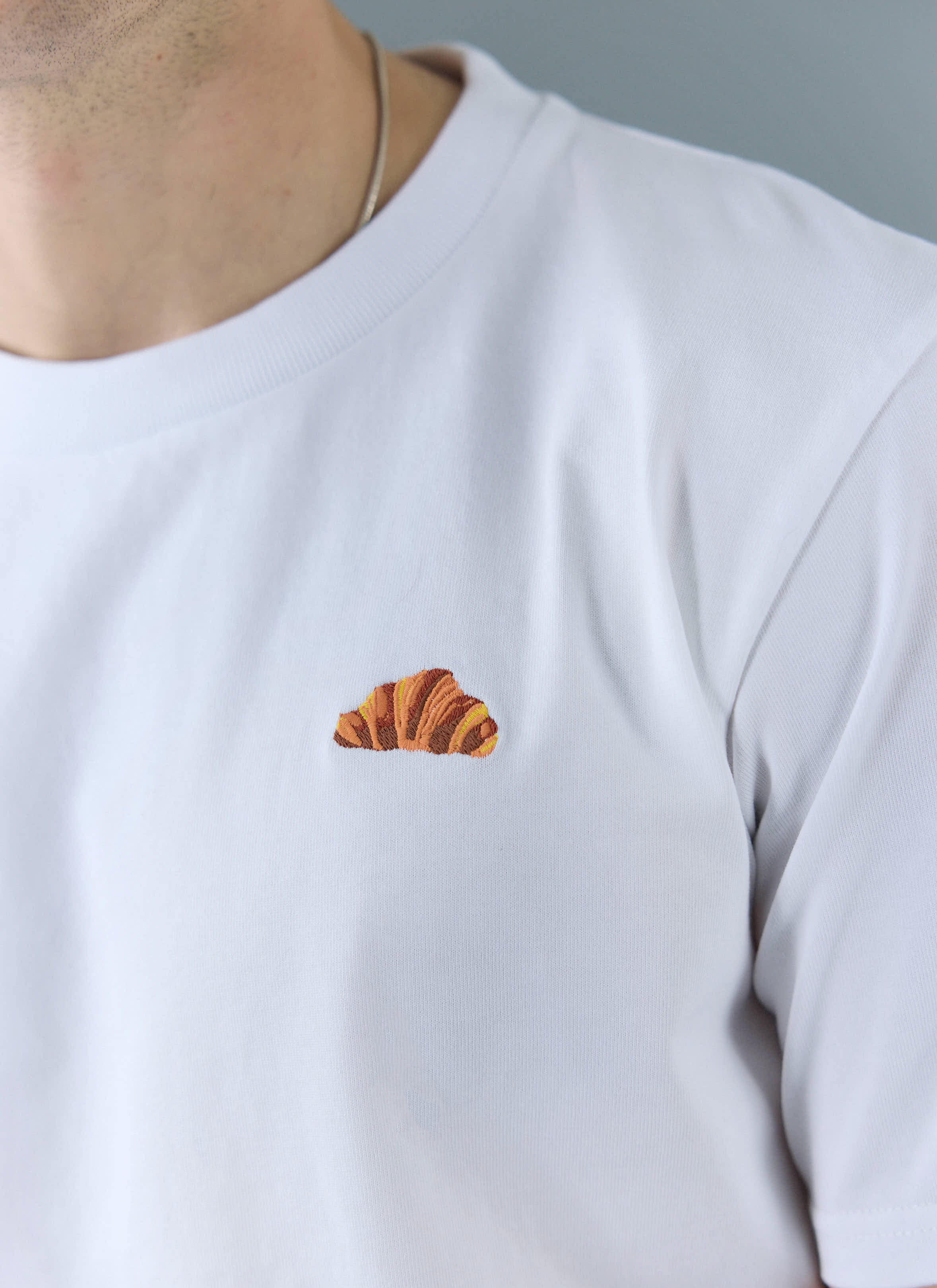 A close up shot of the embroidered croissant detail on the chest of the white organic cotton t-shirt by Percival.