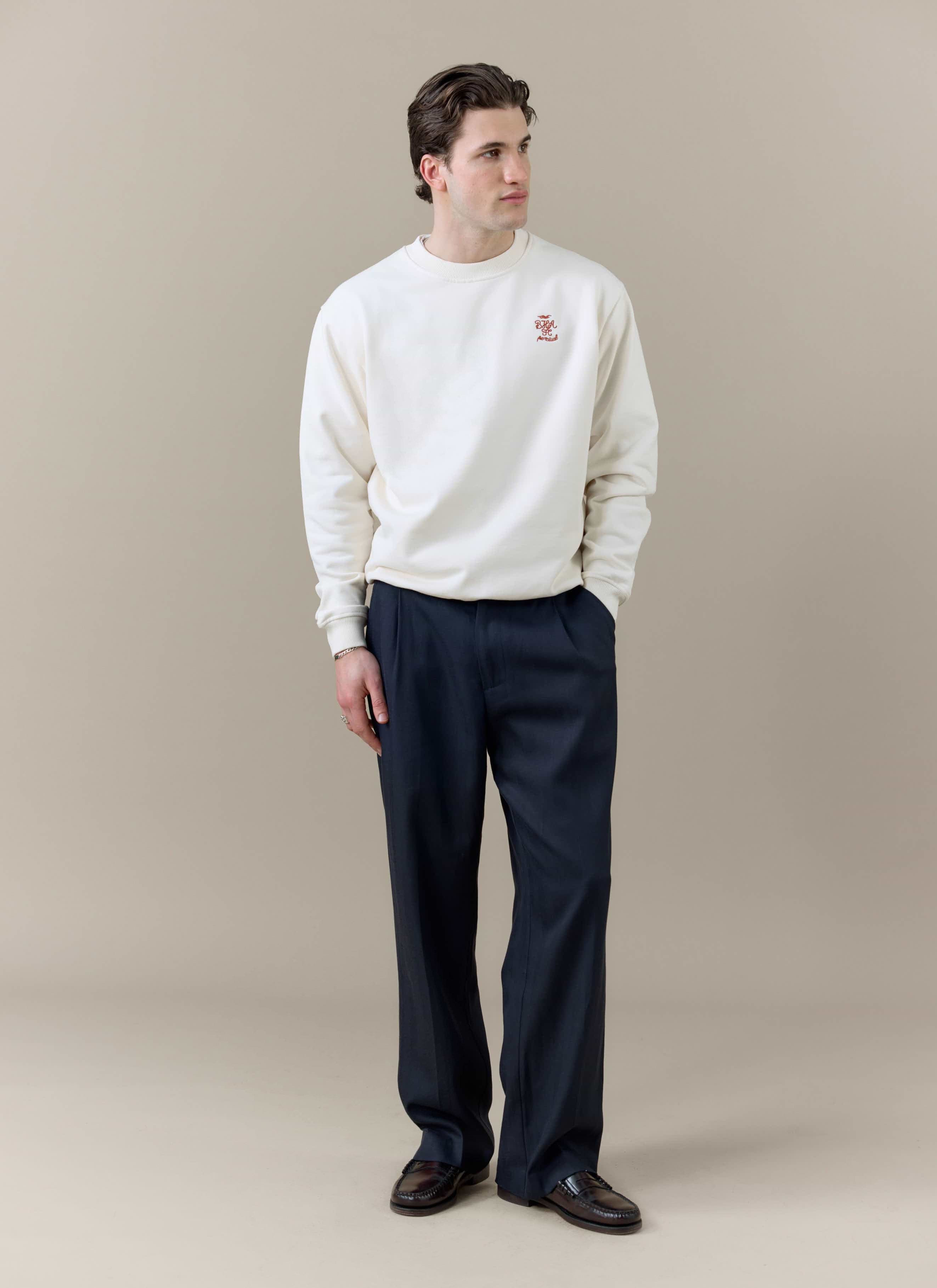 A model wears the cream Percival x Brighton & Hove Albion FC sweatshirt with dark smart trousers against a neutral backdrop.