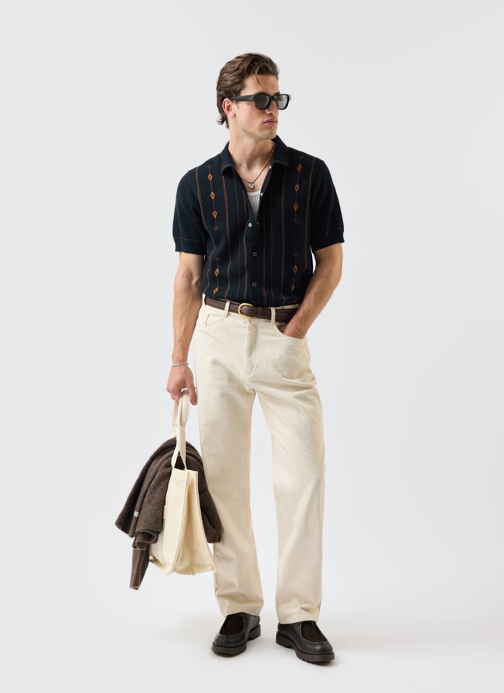 A model wears the Percival Folio Knitted Shirt in dark navy with cream trousers and sunglasses, holding a tote bag.