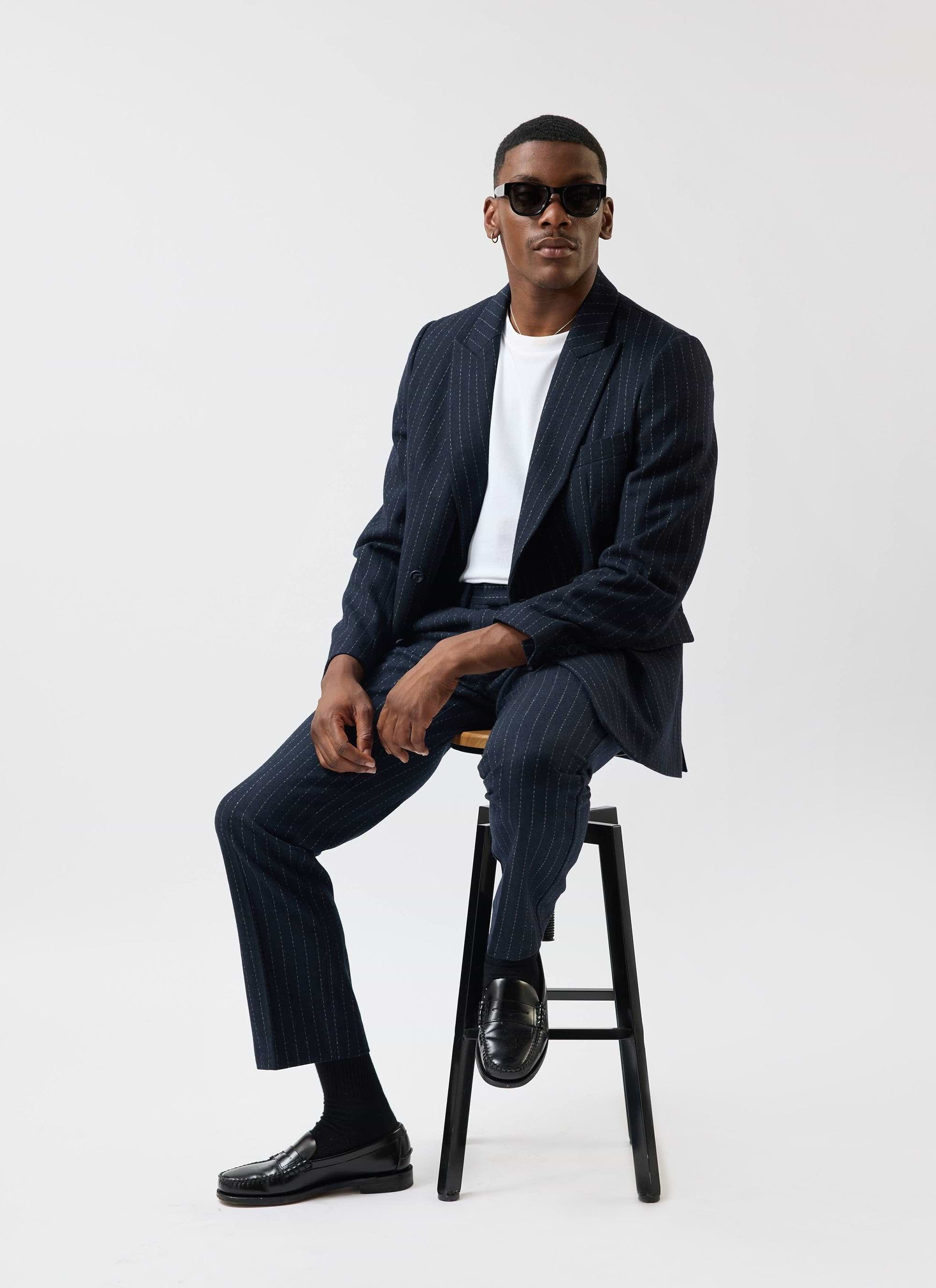 A model sits on a stool wearing the Percival Textured Stripe suit in navy with a white tee, loafers and sunglasses.