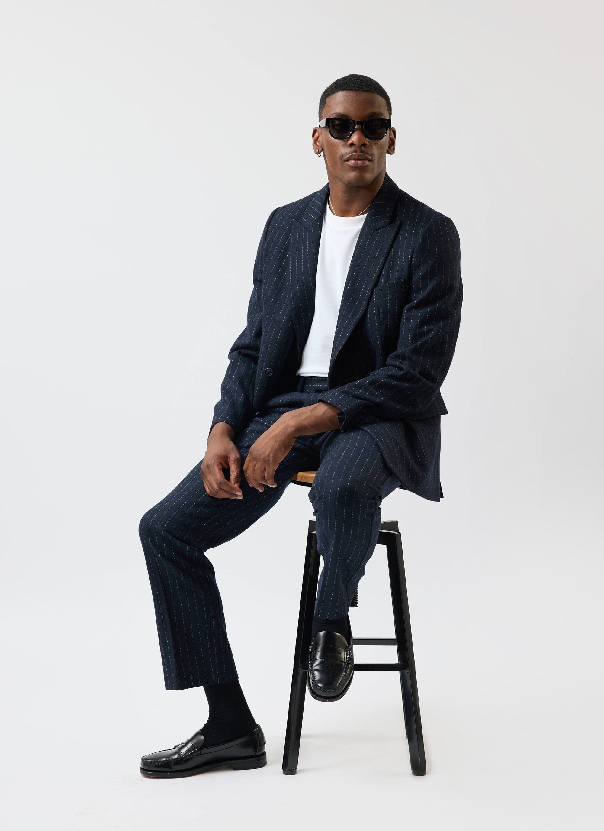 A model sits on a stool wearing the Percival Textured Stripe suit in navy with a white tee, loafers and sunglasses.