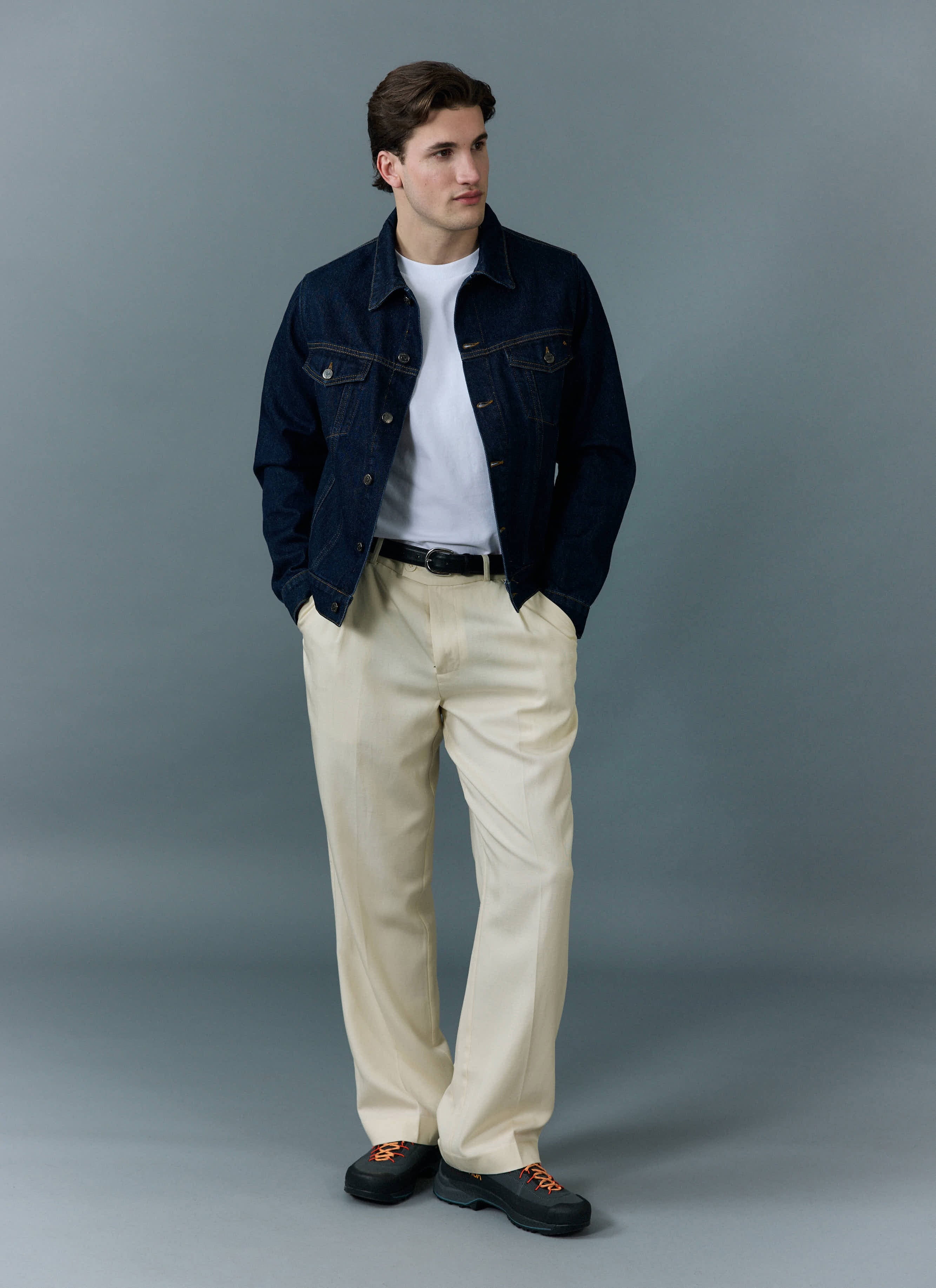 A model wears the Percival Western Trucker Jacket in indigo over a white tee with cream trousers, against a grey background.