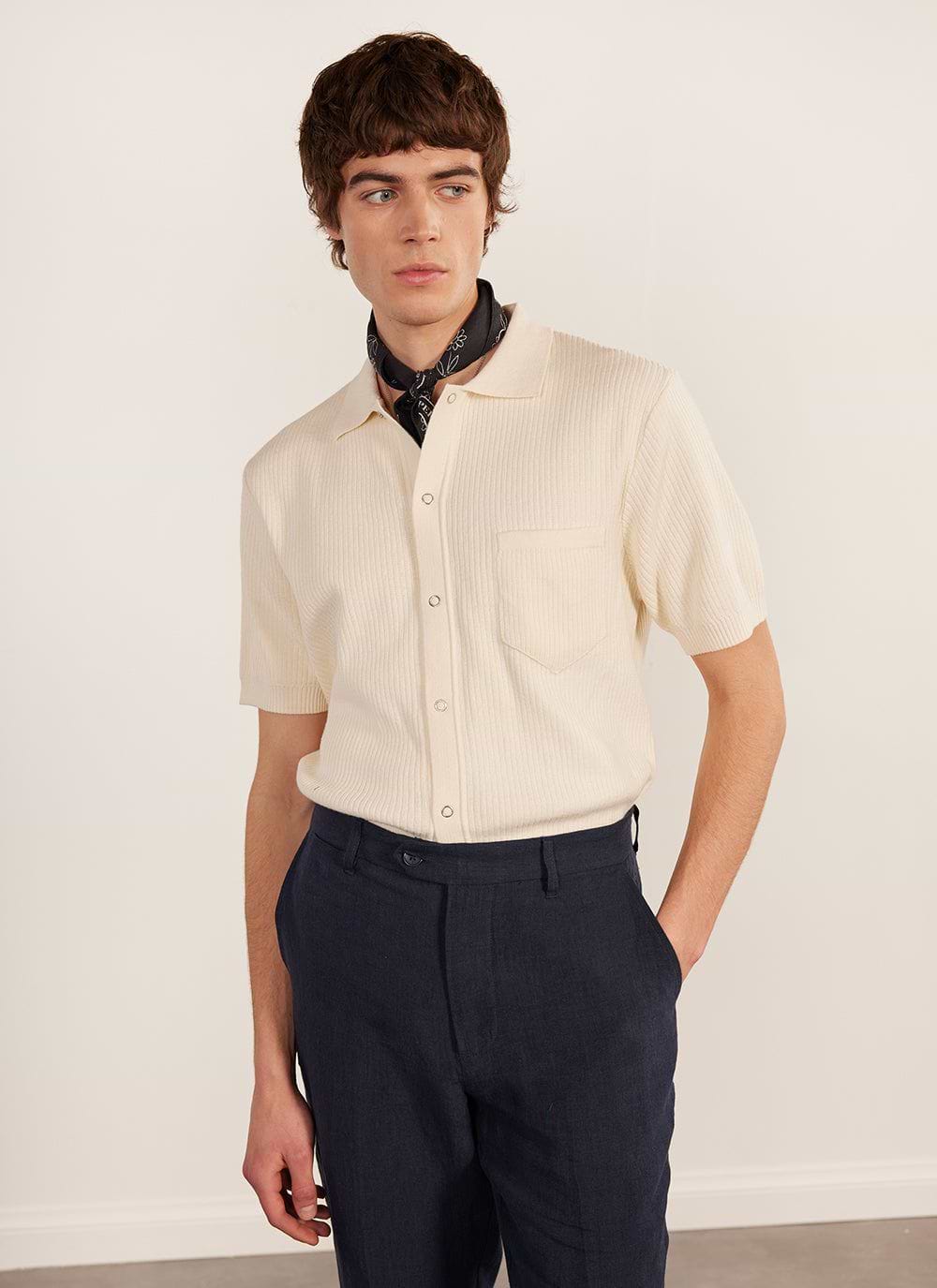 A model wears the Percival Pablo Cuban Shirt in cream, a knitted short sleeve shirt, with dark trousers and a neckerchief.