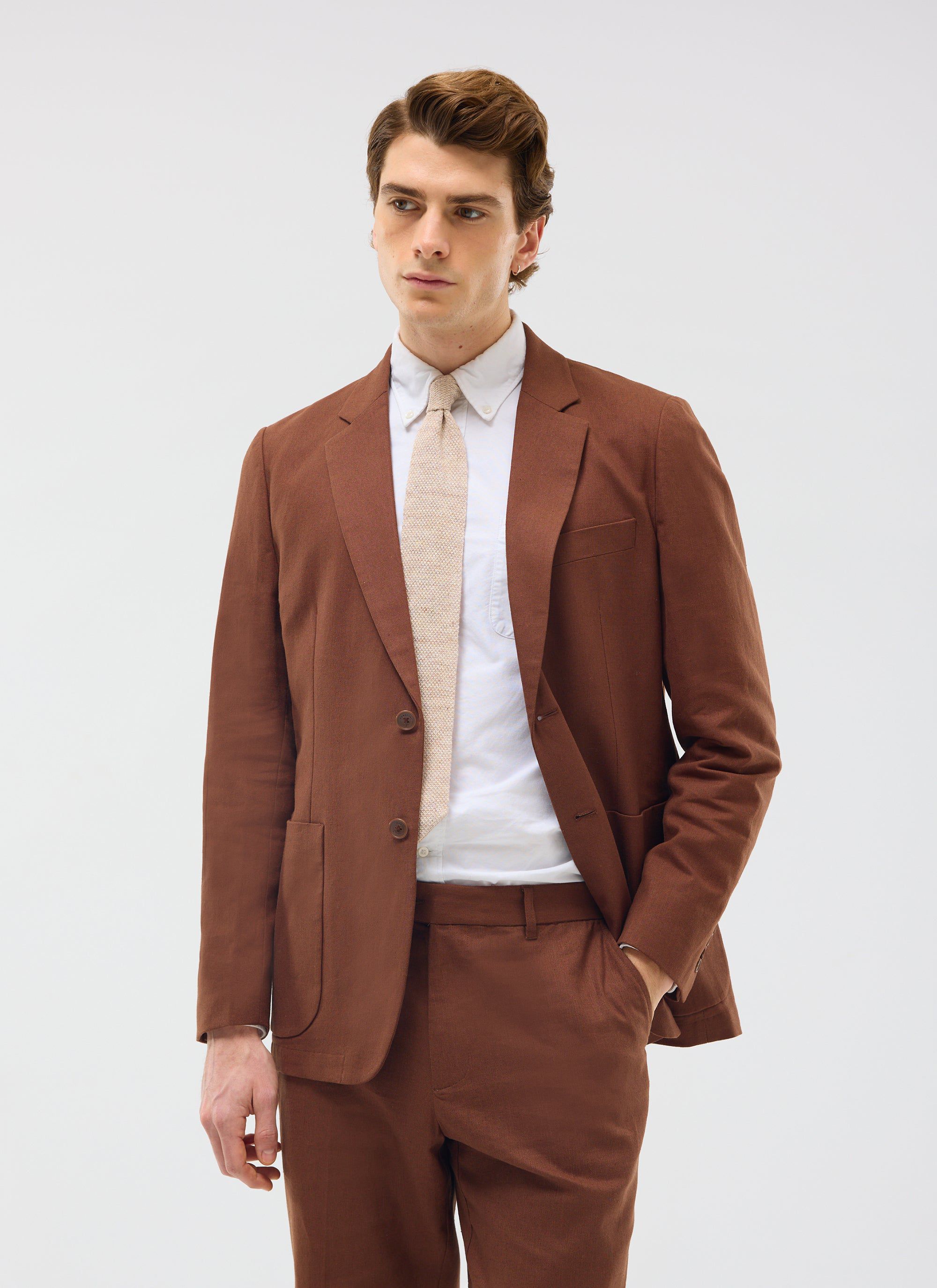 A model wears the Percival Tailored Linen Blazer in brown, styled as a full suit with a white shirt and a light tie.