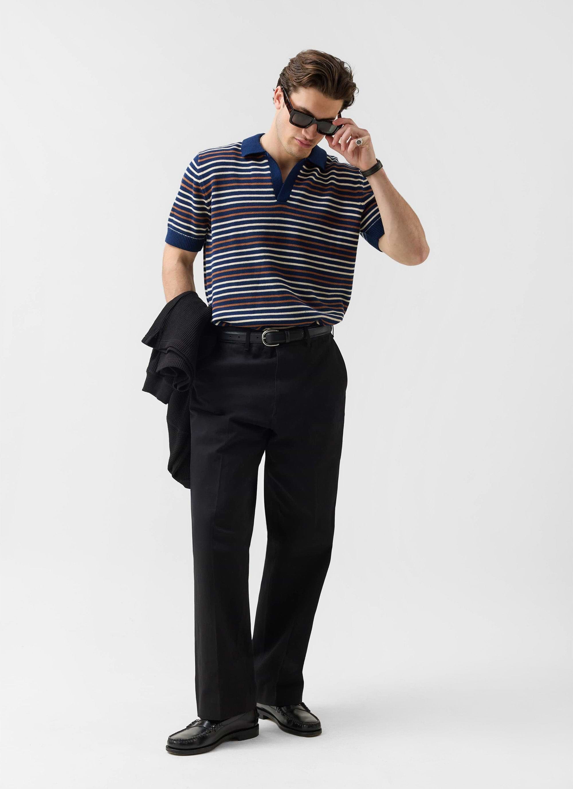 A model wears the Percival Douglas Boxy Knitted Polo in navy stripe, styled with black trousers and sunglasses.