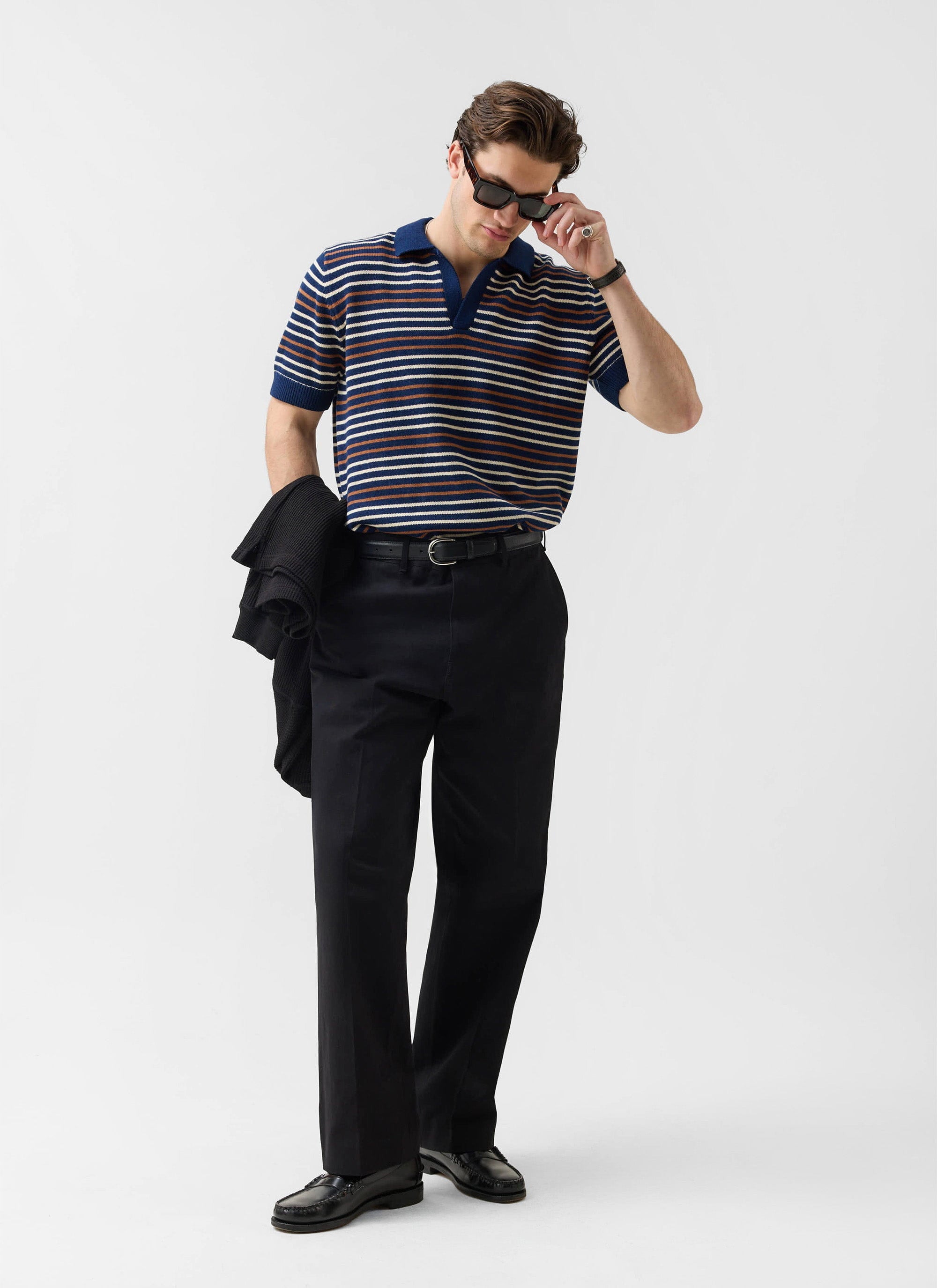 A model wears the Percival Douglas Boxy Knitted Polo in navy stripe, styled with black trousers and sunglasses.