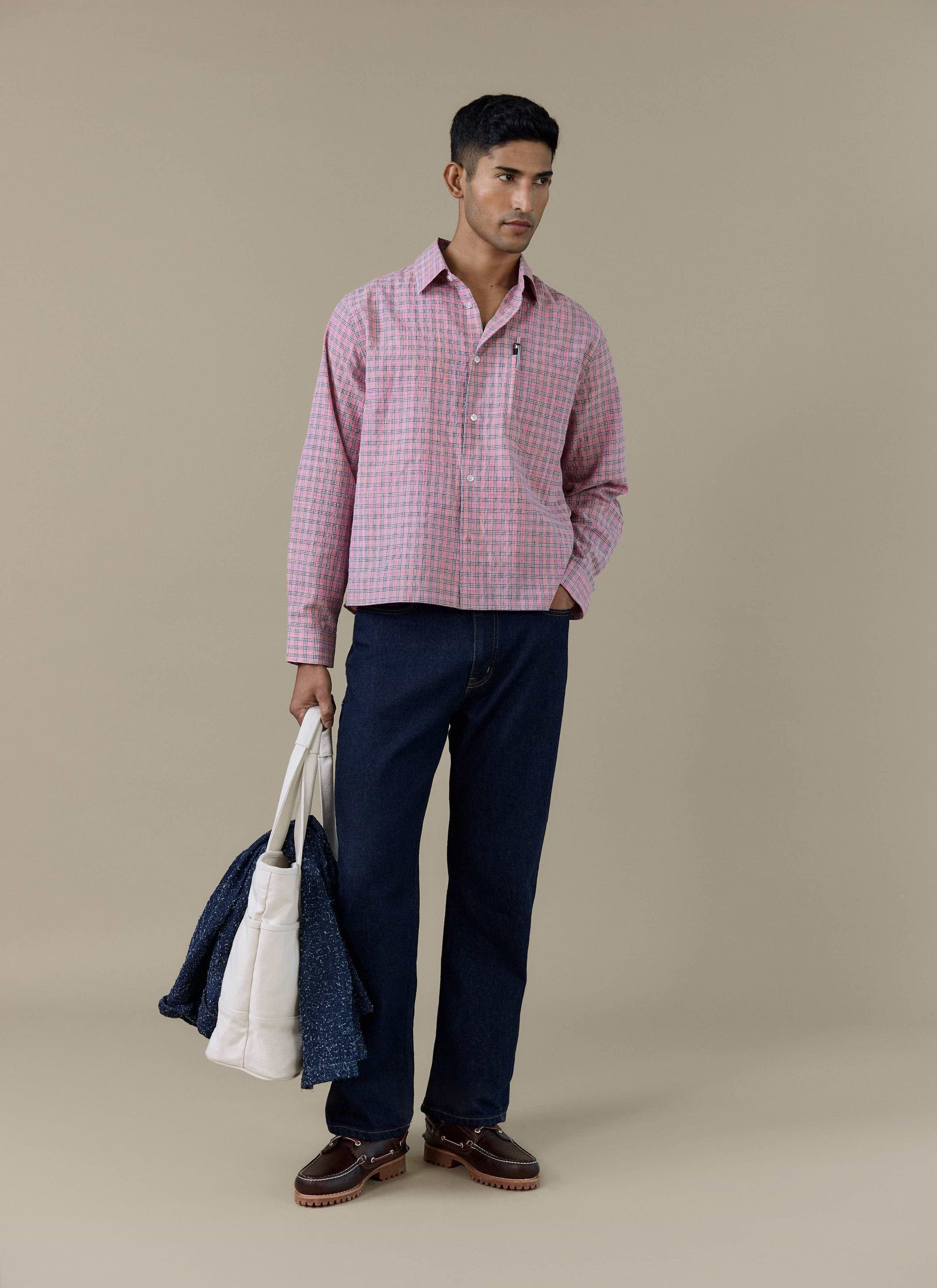 A model wearing the pink Hinsley Checked Boxy Shirt by Percival, styled with dark blue trousers and a white tote bag.