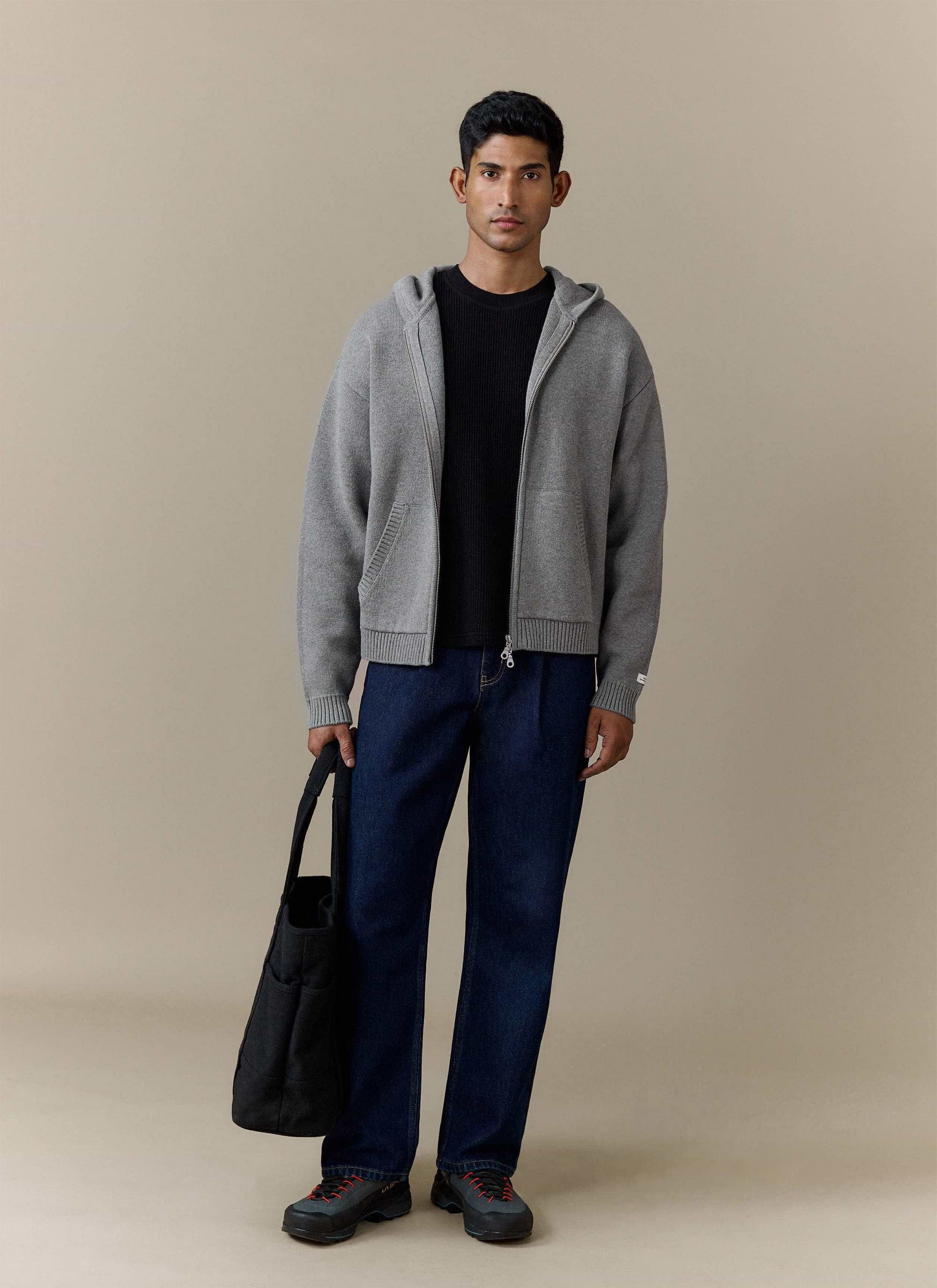 Full shot of a model wearing the Percival Chronicle Knitted Hoodie in grey, layered over a black tee with dark denim jeans.