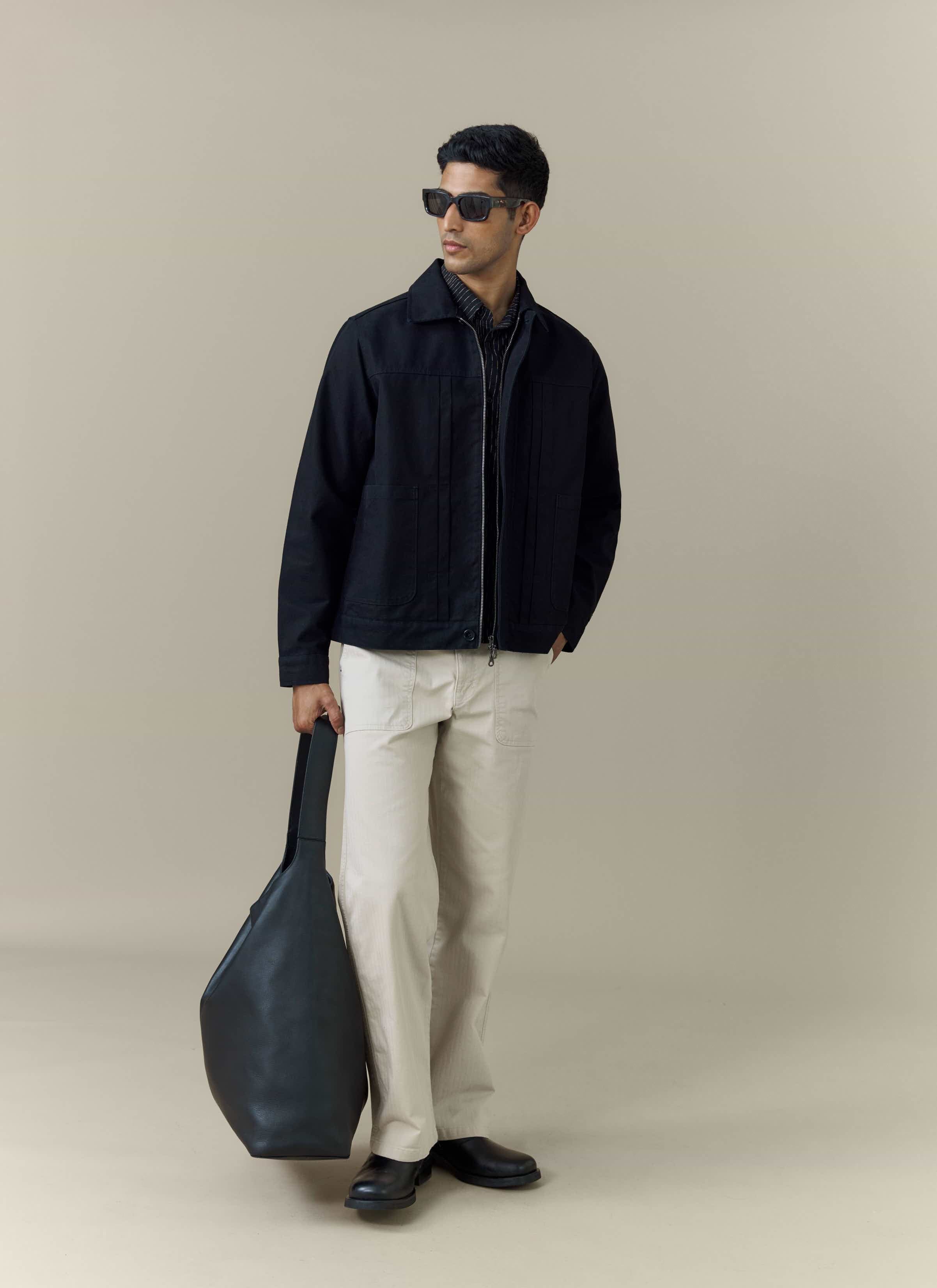 A model wears the black cotton Ernst Jacket from Percival, styled with cream trousers, sunglasses and a black tote bag.