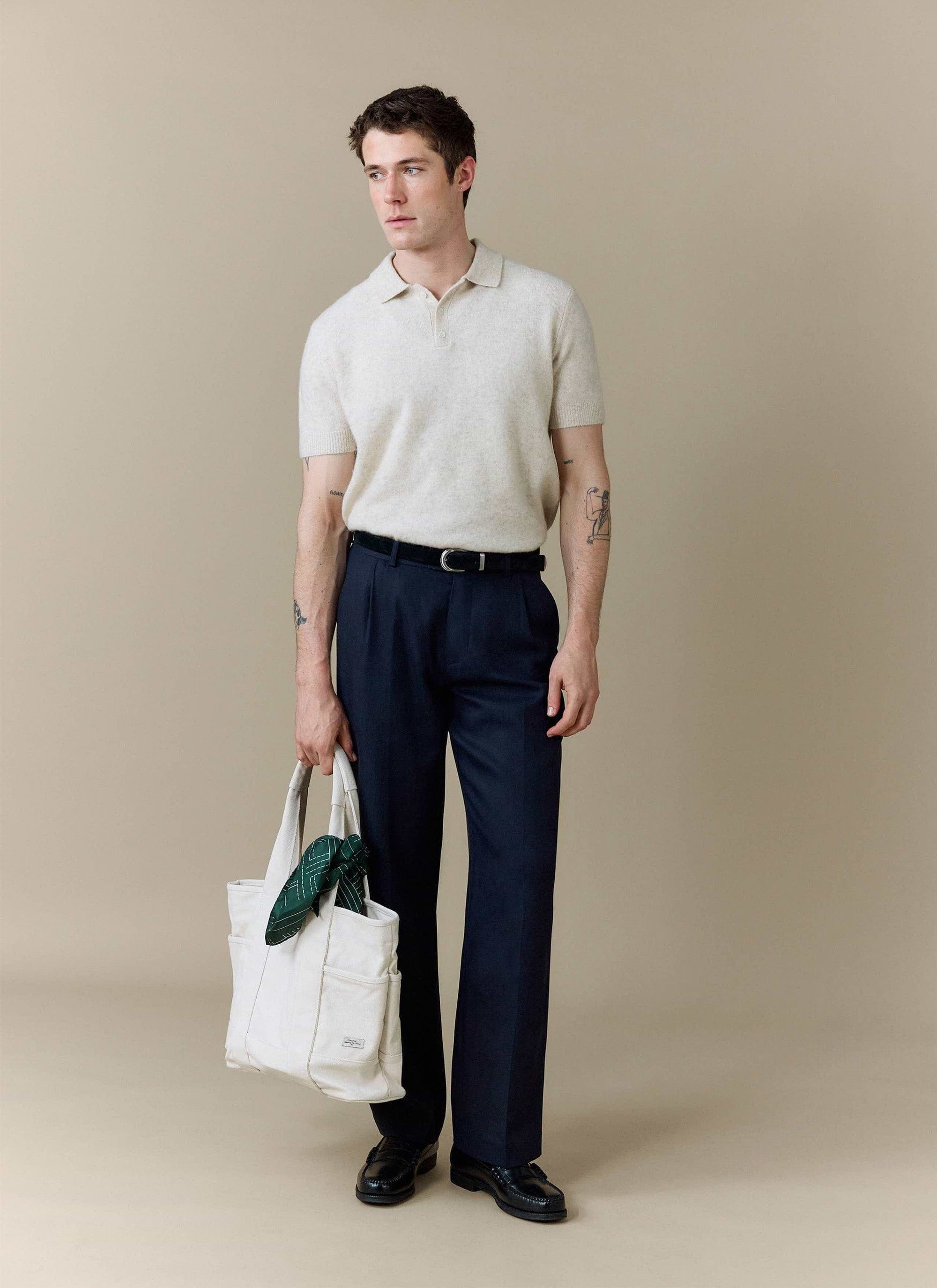 A model wears the Percival Collins Boxy Knitted Polo in ecru, styled with smart navy trousers and a white tote bag.