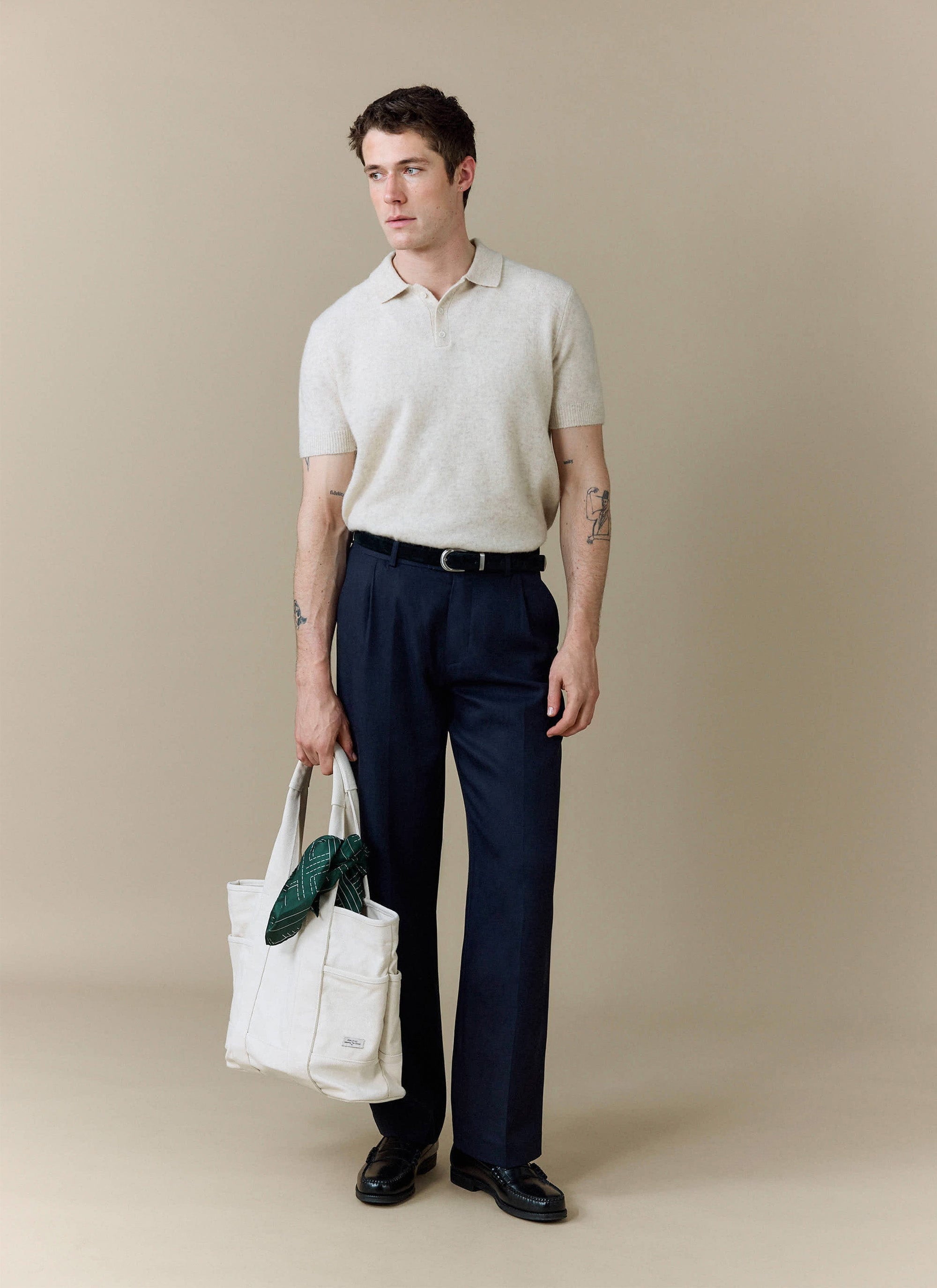 A model wears the Percival Collins Boxy Knitted Polo in ecru, styled with smart navy trousers and a white tote bag.