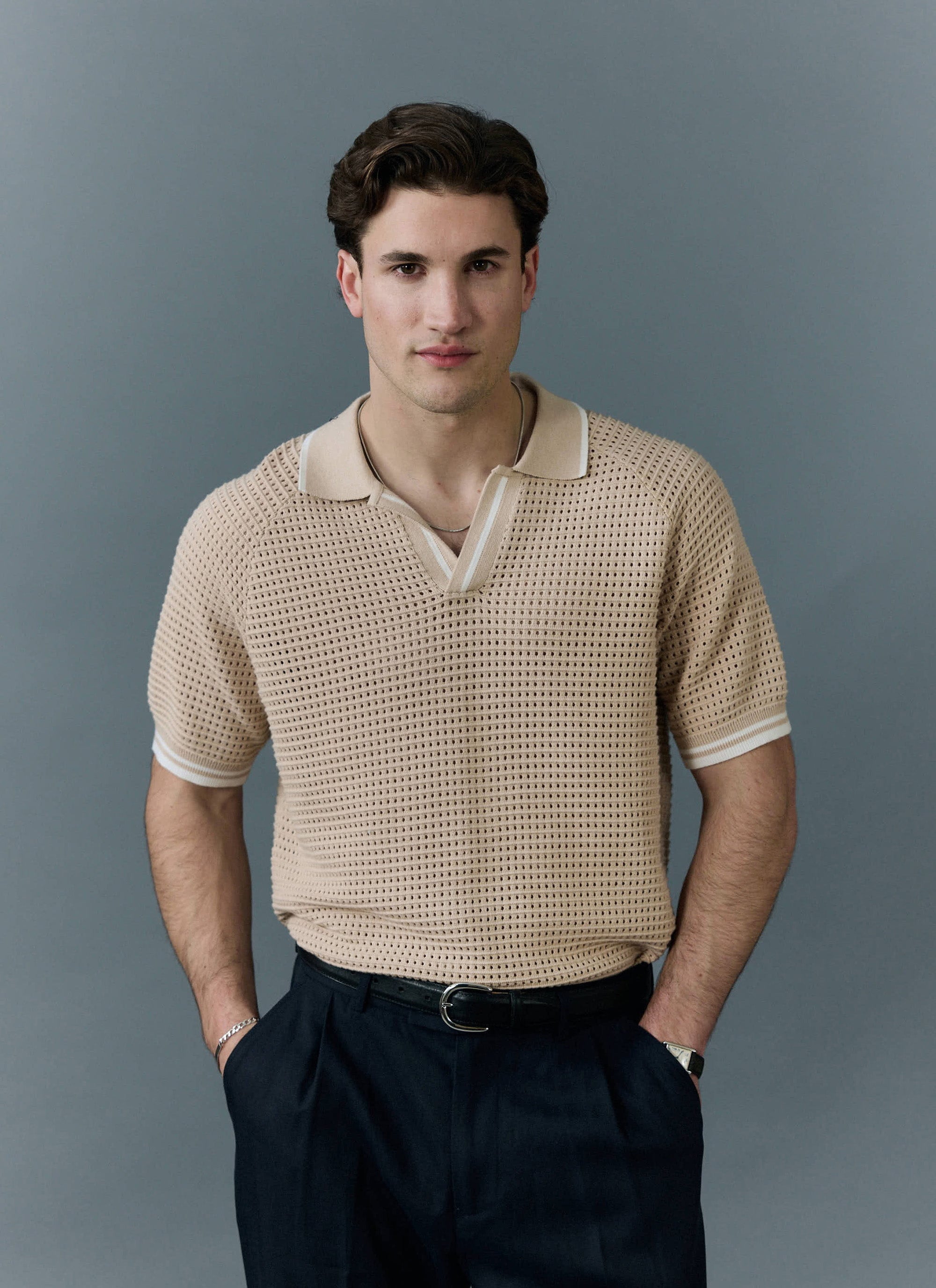 A model wears the Percival Tennis Boxy Knitted Polo in ecru, a cotton shirt with a perforated knit, on a grey background.