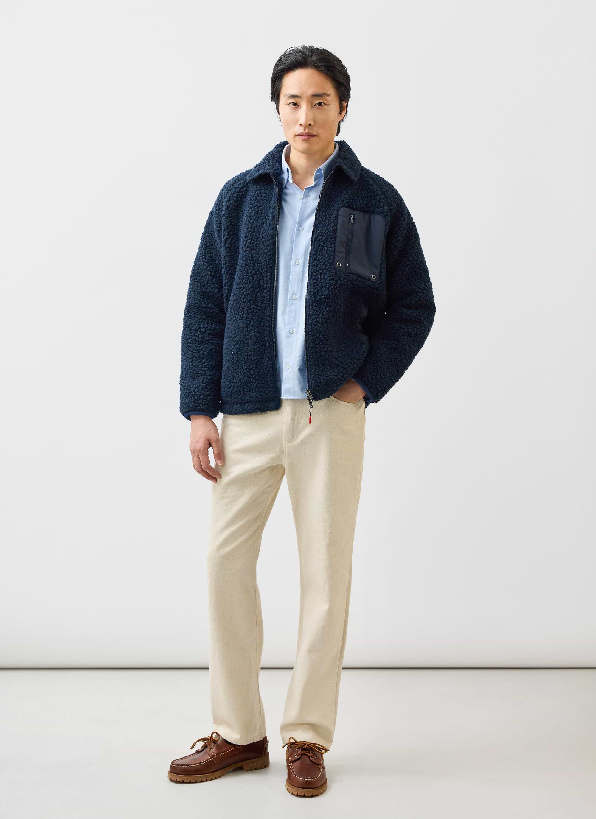 A model wears the navy wool fleece jacket from Percival with a light blue shirt, cream trousers, and brown shoes.