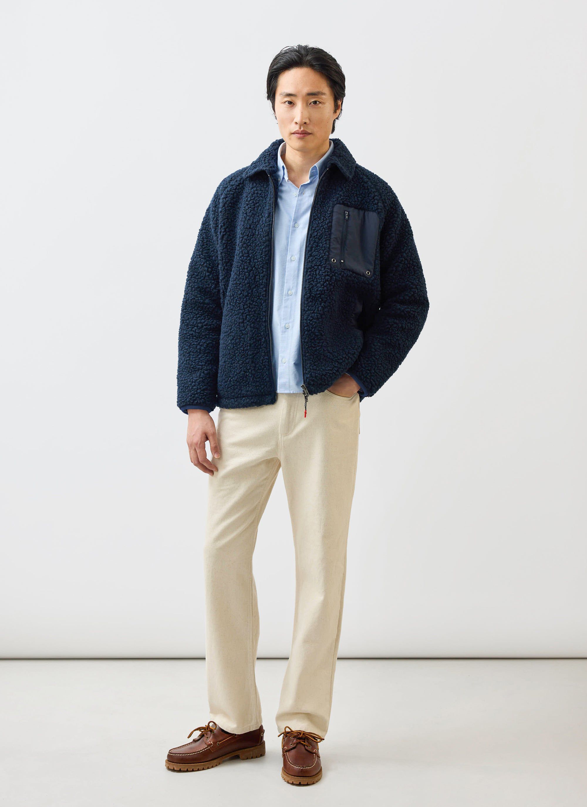 A model wears the navy wool fleece jacket from Percival with a light blue shirt, cream trousers, and brown shoes.
