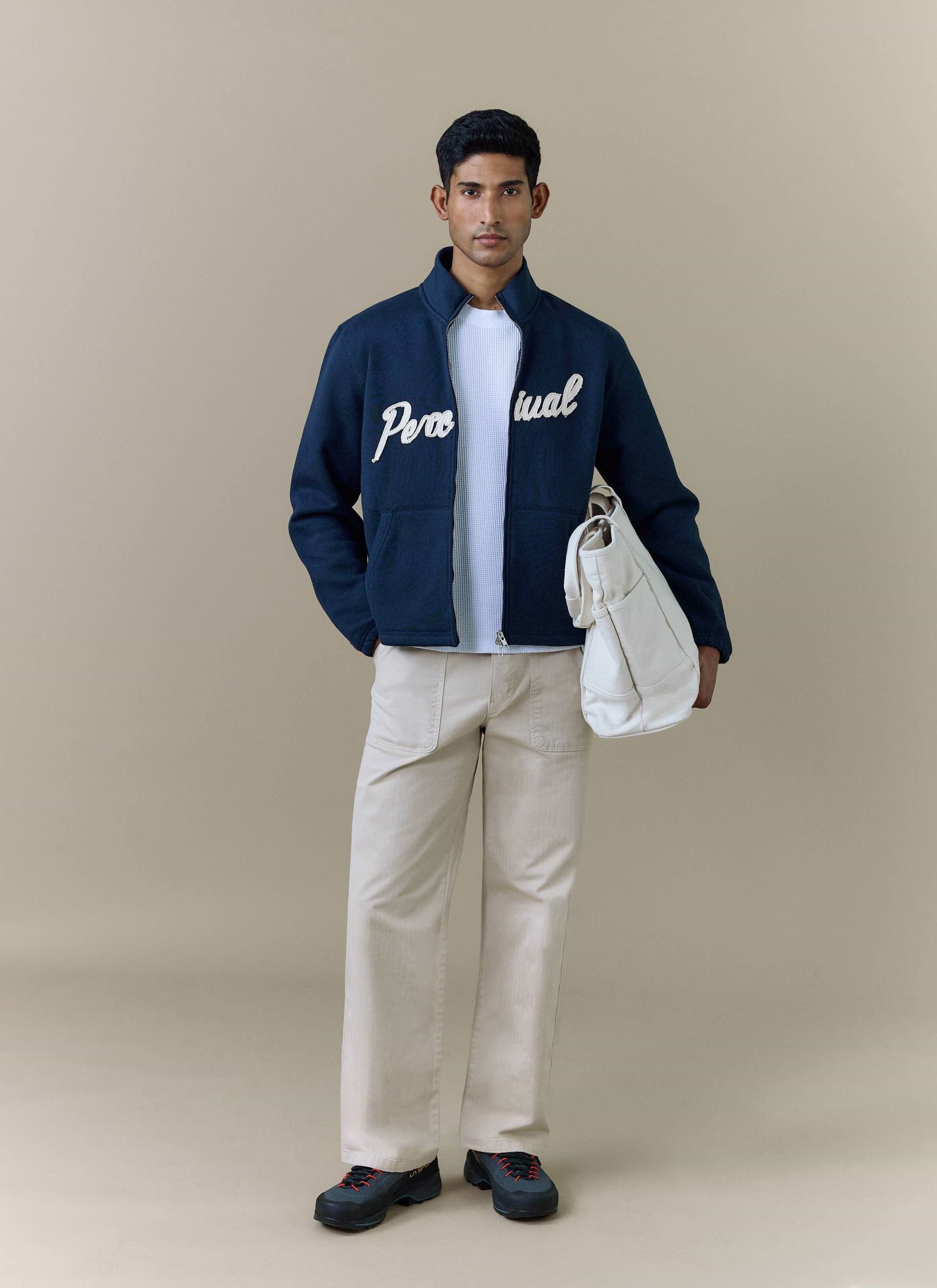 A model wears the navy Curtis Boxy Harrington jacket from Percival, styled with cream trousers and holding a white tote bag.