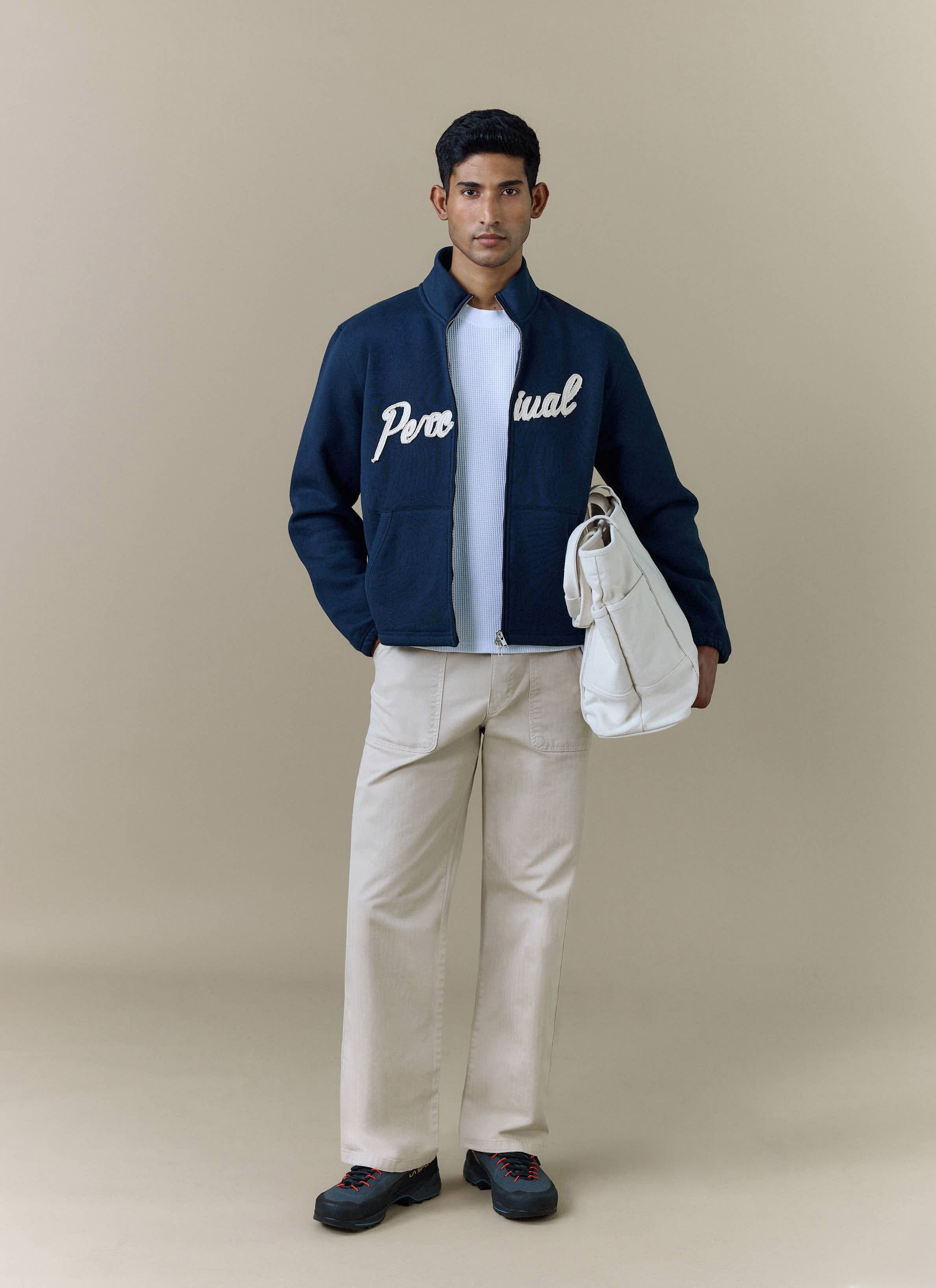 A model wears the navy Curtis Boxy Harrington jacket from Percival, styled with cream trousers and holding a white tote bag.