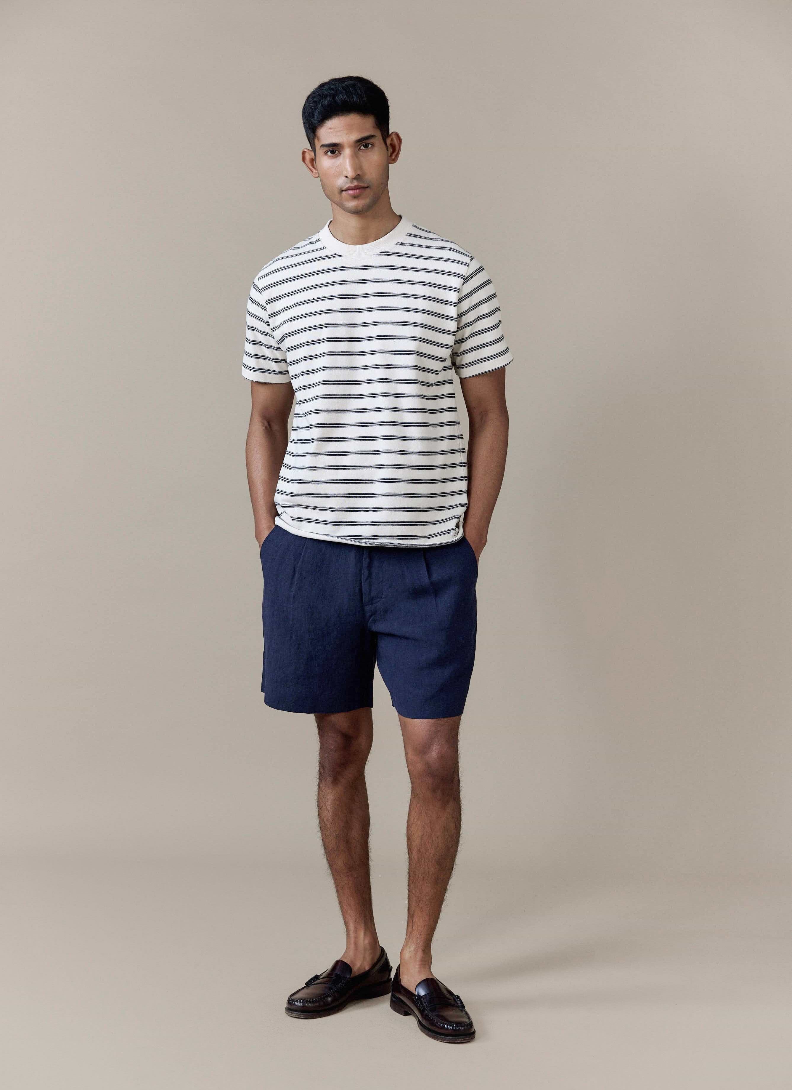 A model wears navy pleated linen shorts by Percival with a striped t-shirt, featuring a tailored fit and front pleats.